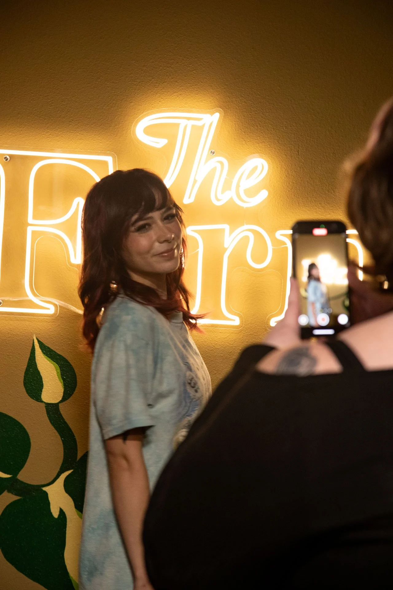 A woman with reddish-brown hair posing for a photo in front of a yellow wall with a neon sign that says 'The Fern' and some green decorative elements. Someone is taking her picture with a smartphone.
