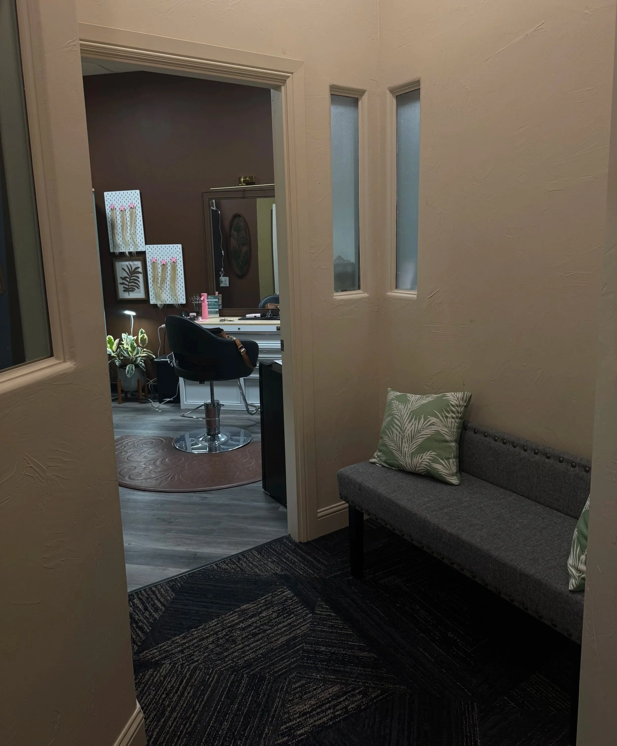 View of an office space seen through a doorway with three small narrow windows on the right. The office has a dark wall, a white desk, a black office chair, and a few decorations, plants, and a lamp. The hallway has a dark carpet and a gray upholstered bench with a patterned pillow.