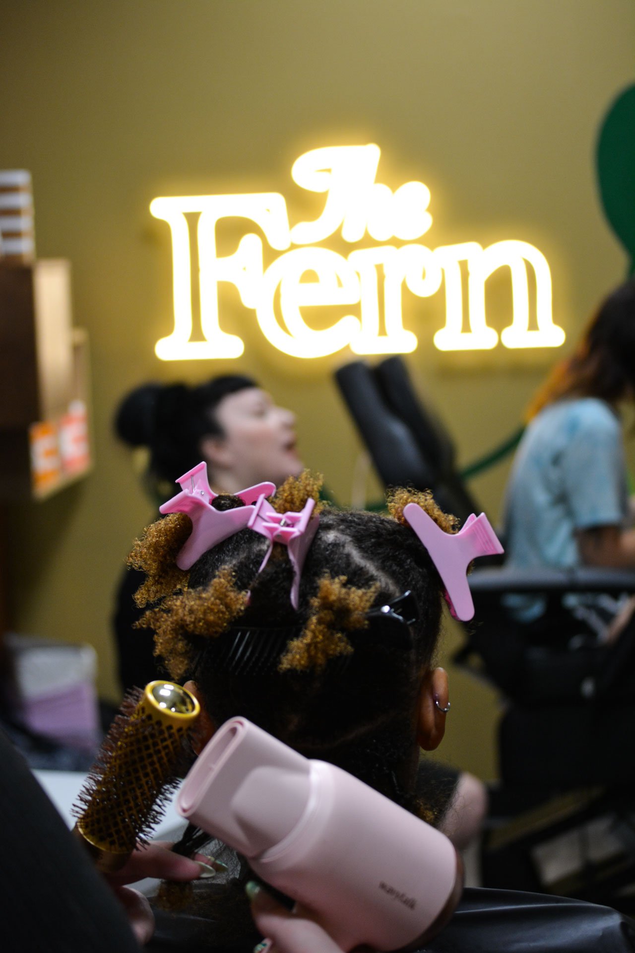 Person with curly hair and pink hair clips getting hair styled with a round brush and hairdryer in a salon, background features a neon sign that says "The Fern"