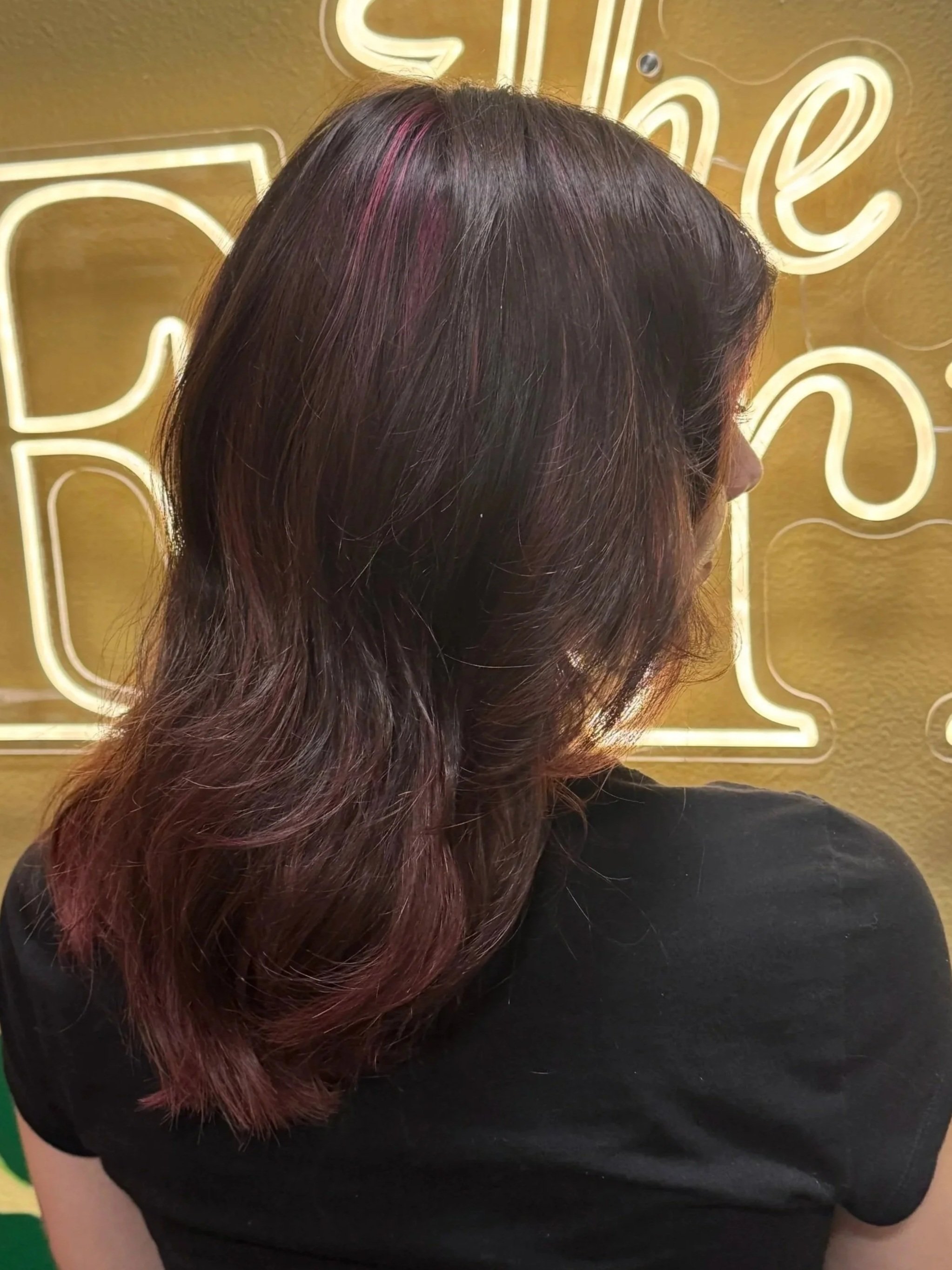 A woman with long brown hair that has pink highlights, wearing a black shirt, standing in front of a yellow wall with neon lights.