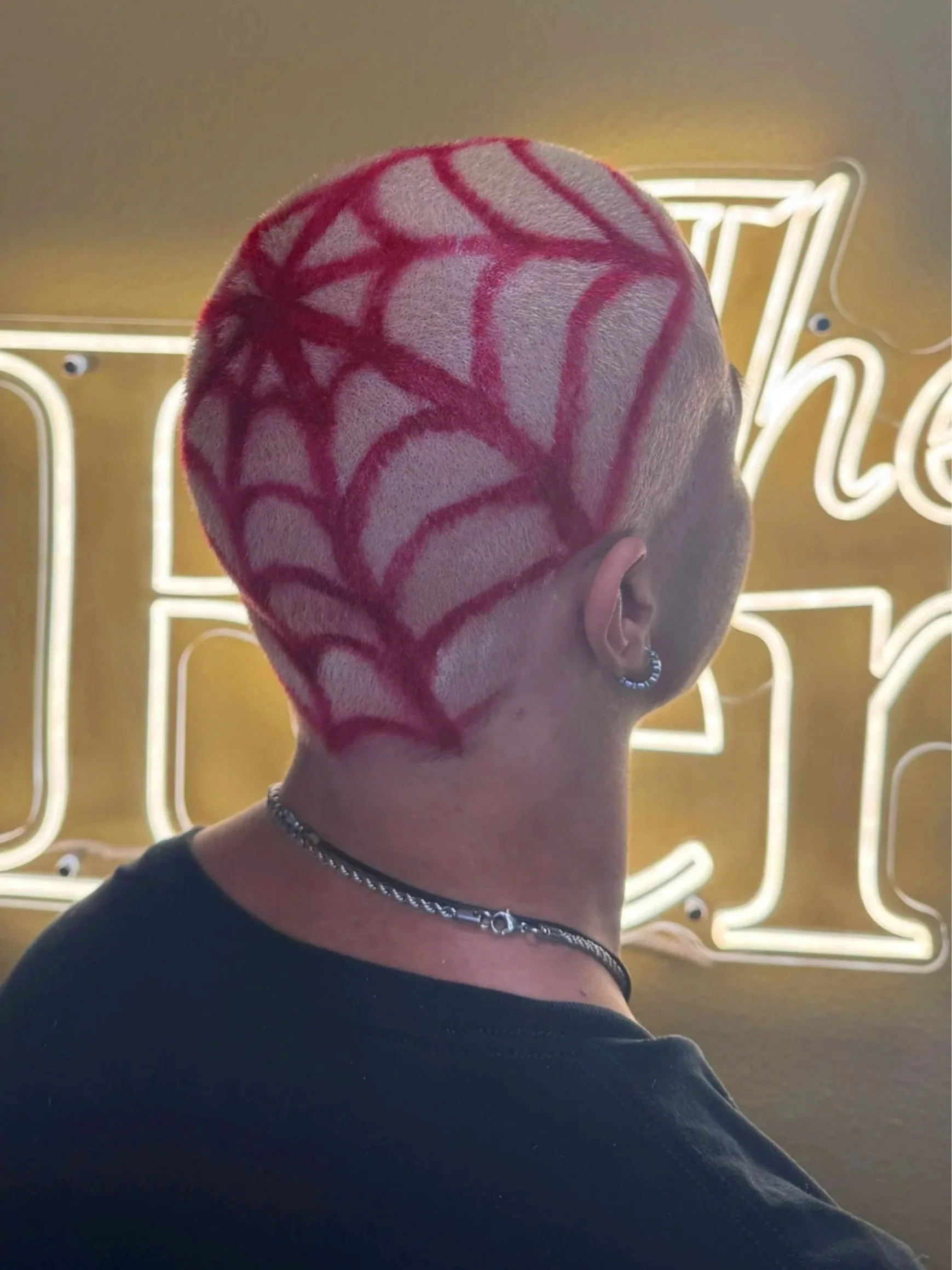 Back of a person's head with a red spider web pattern design on short hair, wearing a black top, necklace, and silver hoop earring, with neon yellow signage in the background.