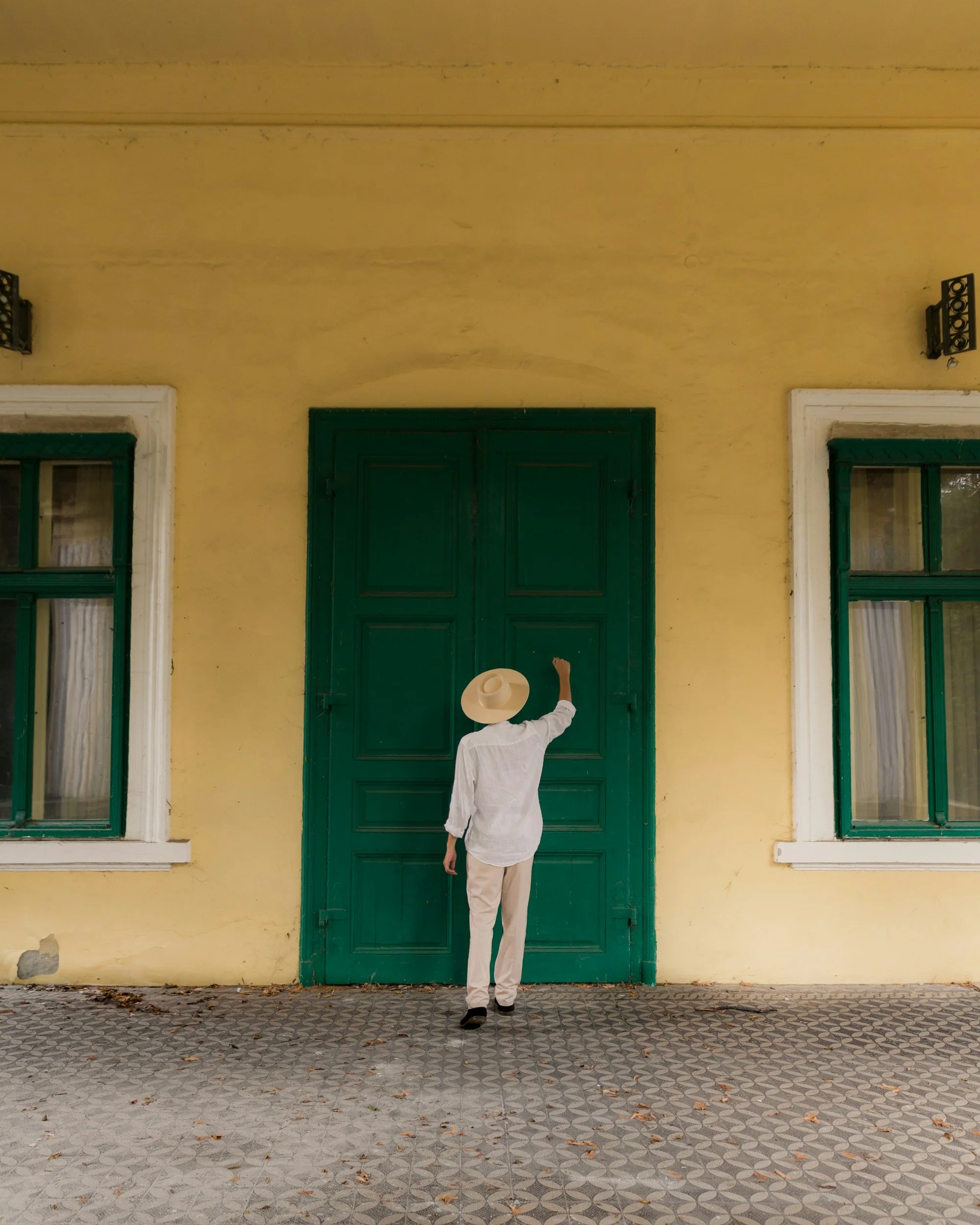 A person dressed in white with a wide-brimmed hat is standing in front of a large green door on a yellow wall with two narrow windows on either side, and cobblestone ground with scattered leaves.
