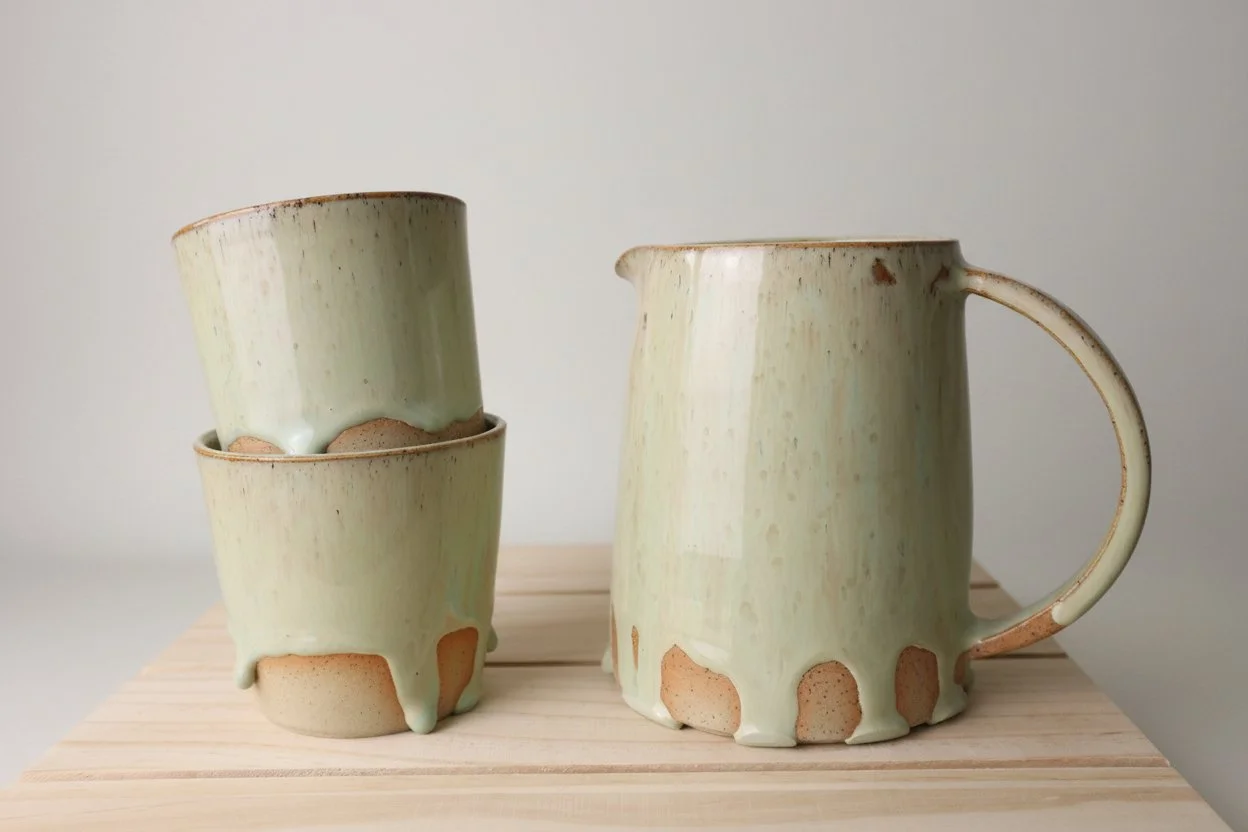 Cream-colored ceramic mug and two matching cups with drip glaze, placed on a light wooden surface.