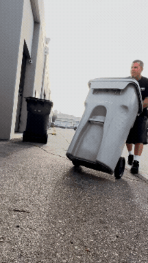 A person pushing a large gray trash can on wheels outside a building.