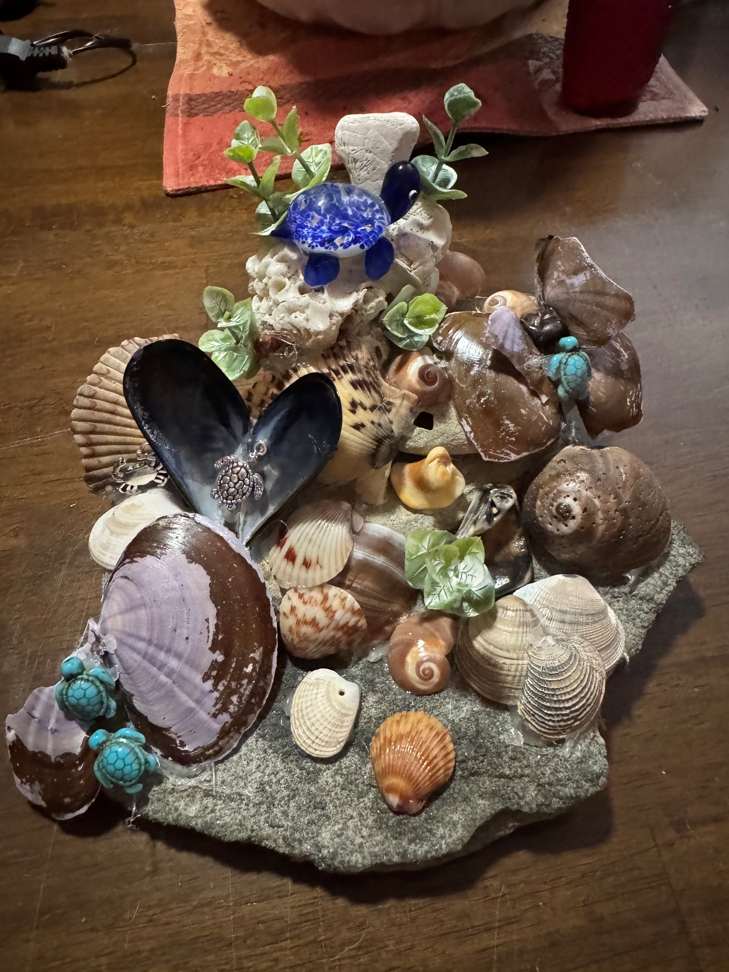 A collection of various seashells, coral, a ceramic blue turtle, a black and white shell, a turquoise bracelet, and a small green leafy plant arranged on a piece of rock.