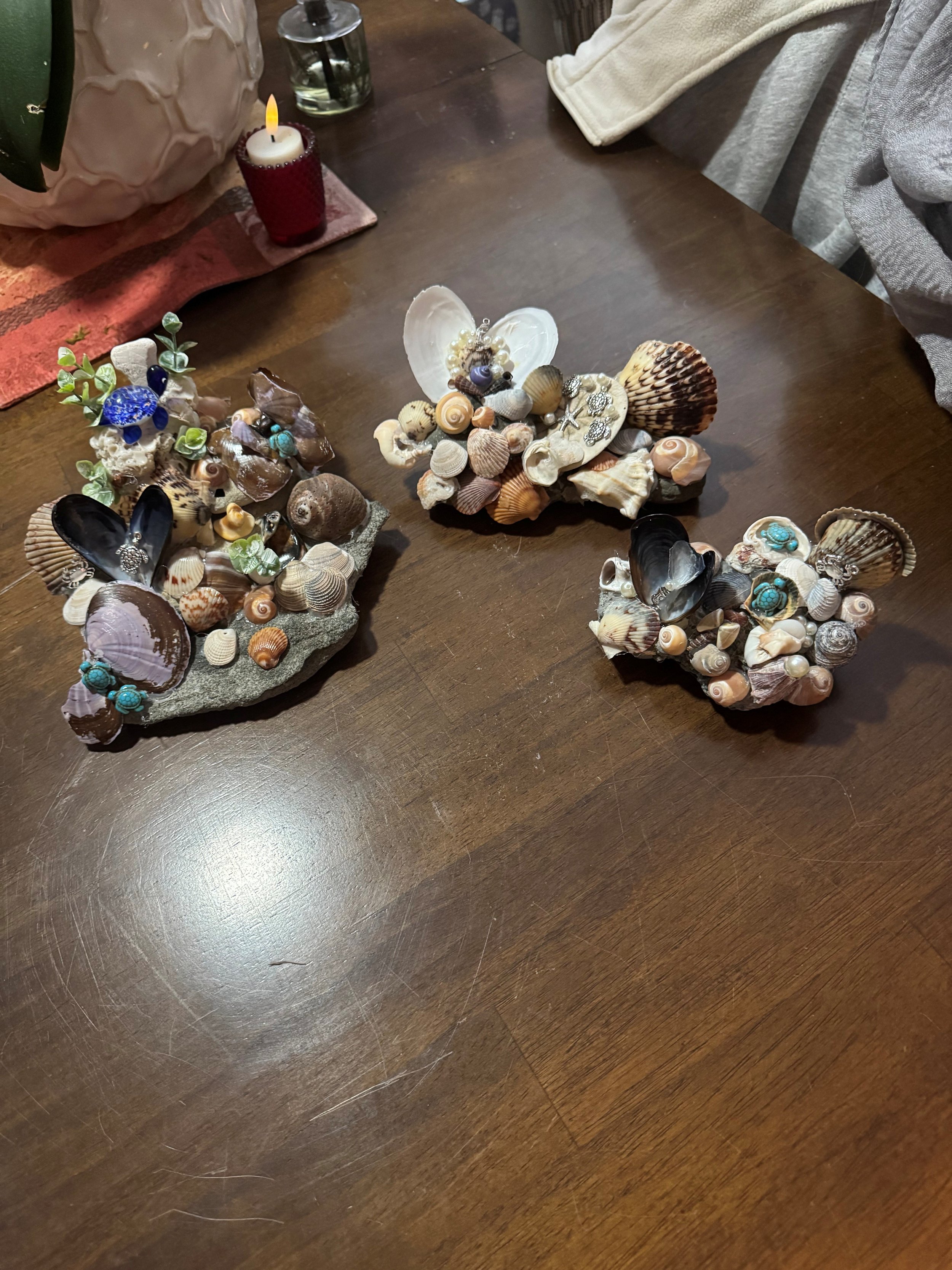 Three coral and shell arrangements with decorative jewelry on a dark wooden table, with a candle and glass of water in the background.