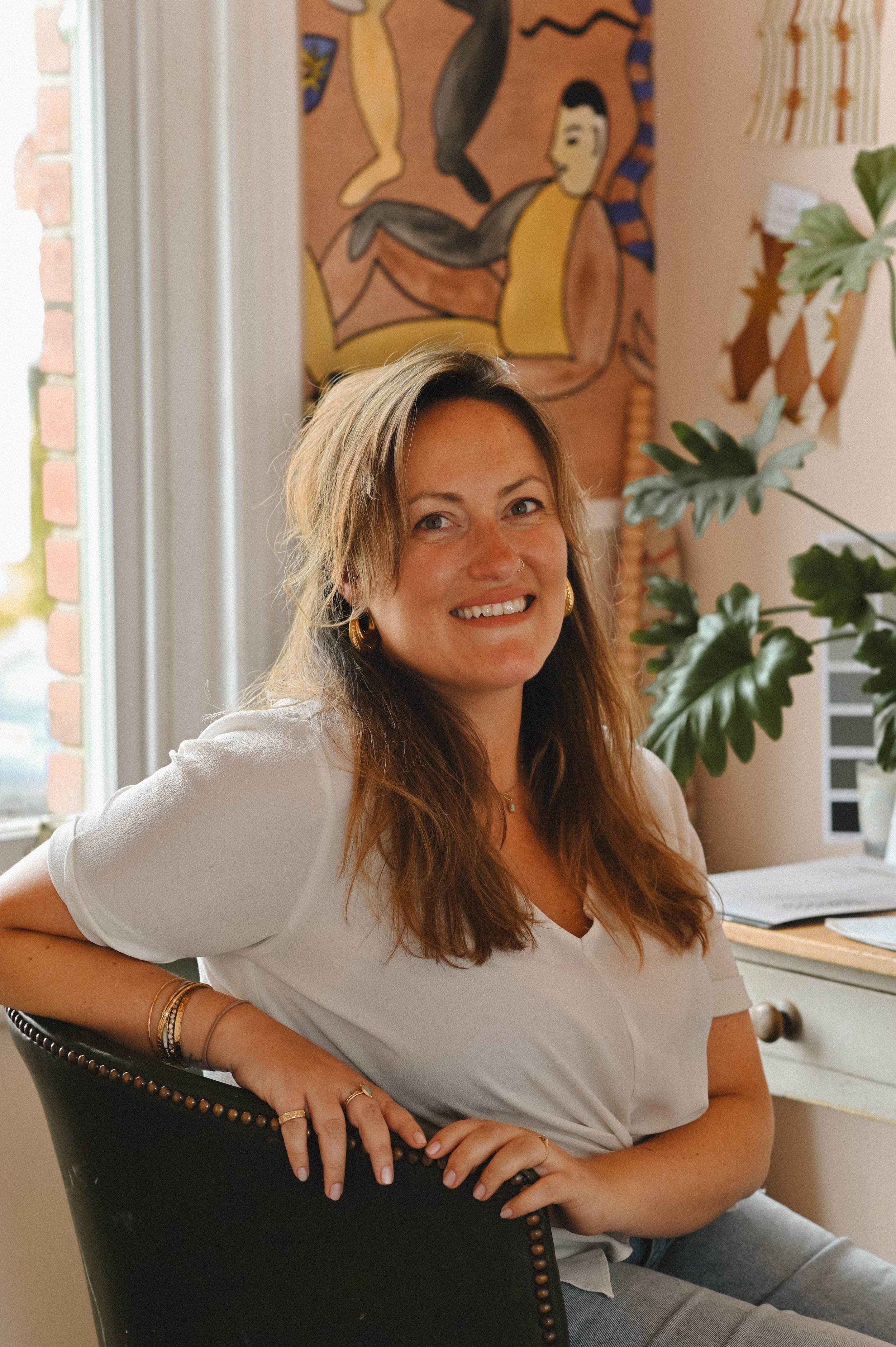 A woman with long wavy hair, fair skin, and wearing a white shirt, smiling while sitting in a black chair with nailhead trim. She has gold jewelry, including bracelets and earrings. The background features a colorful abstract painting, a window with brick window frame, a houseplant, and a desk with papers.