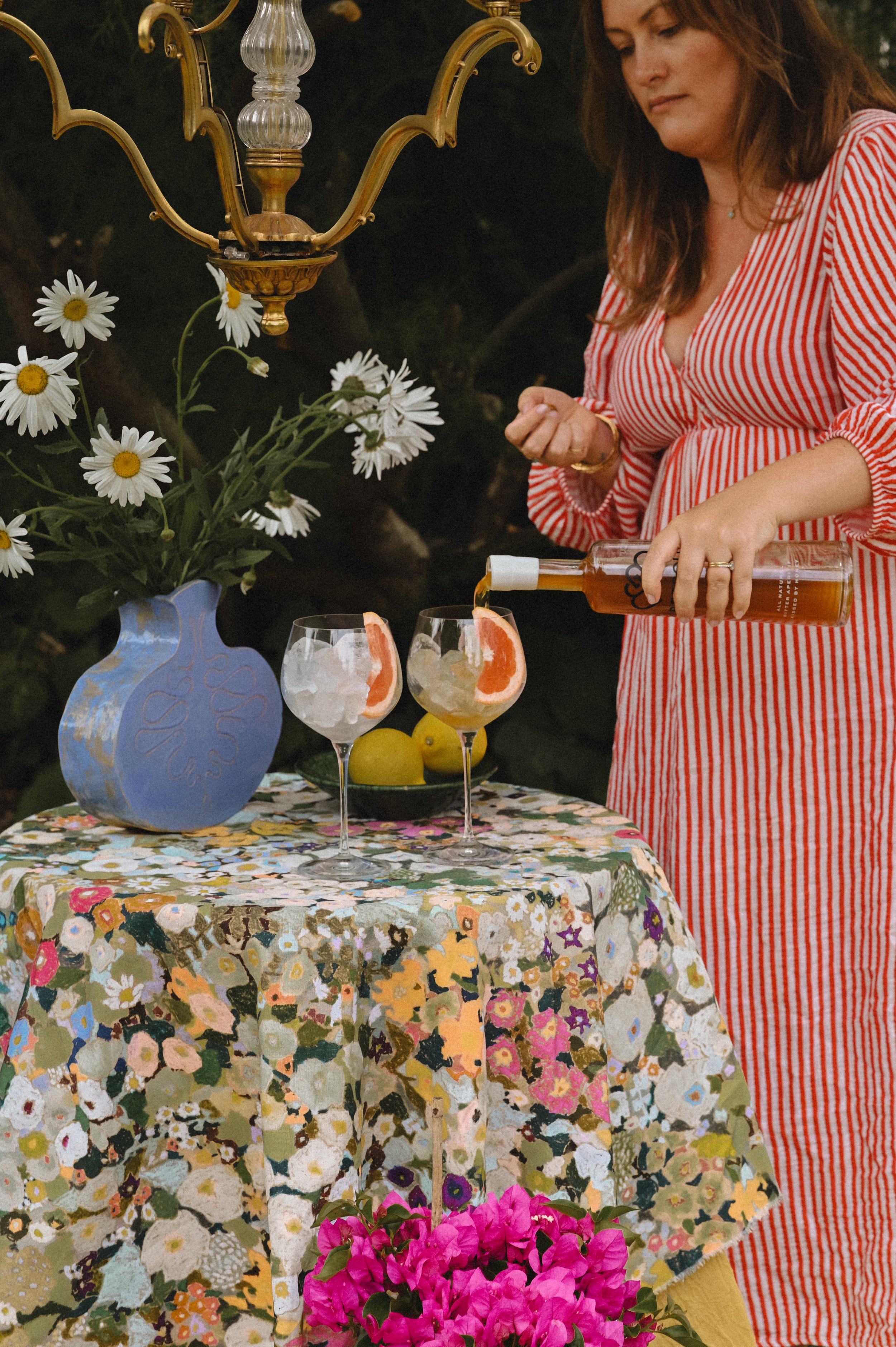 A woman in a red and white striped dress pouring a drink into two glasses on a table with a colorful floral tablecloth. The table has a vase with white daisies, a bowl with lemons, and a bright pink bouquet of flowers. An antique chandelier hangs above.