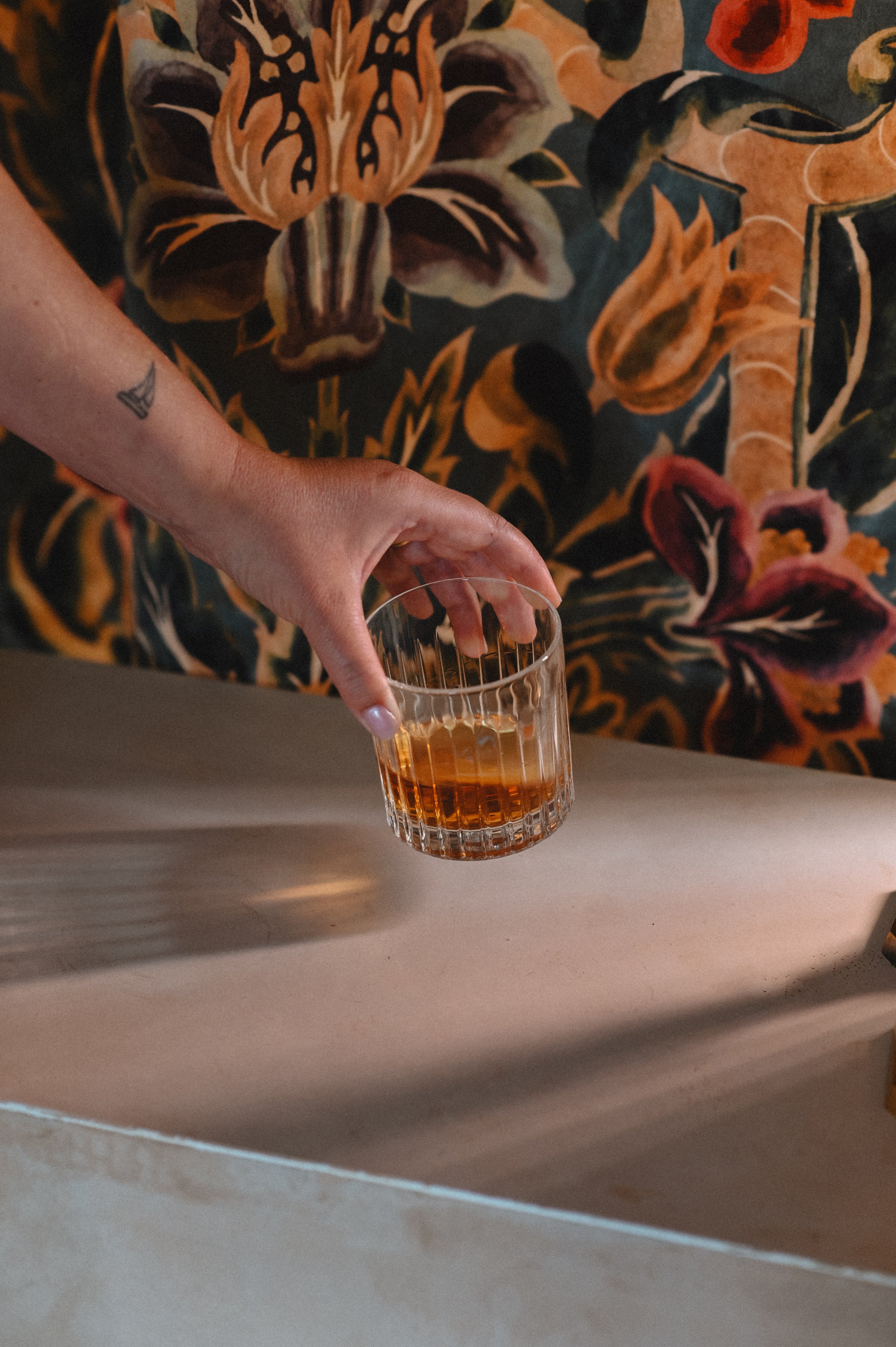 A person wearing a floral patterned shirt holds a glass of amber-colored beverage.