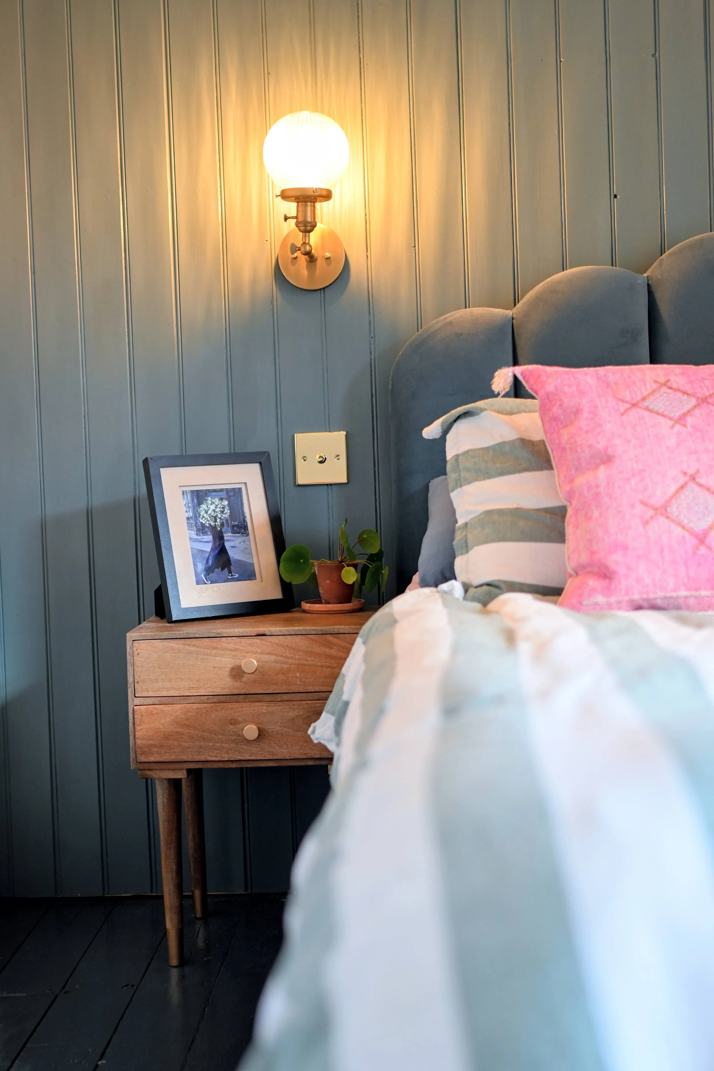 A bedroom corner with a wooden nightstand, a framed photo of a dog, a potted plant, a textured headboard, colorful pillows, a blue wall with vertical paneling, a wall lamp, and a visible electrical outlet.