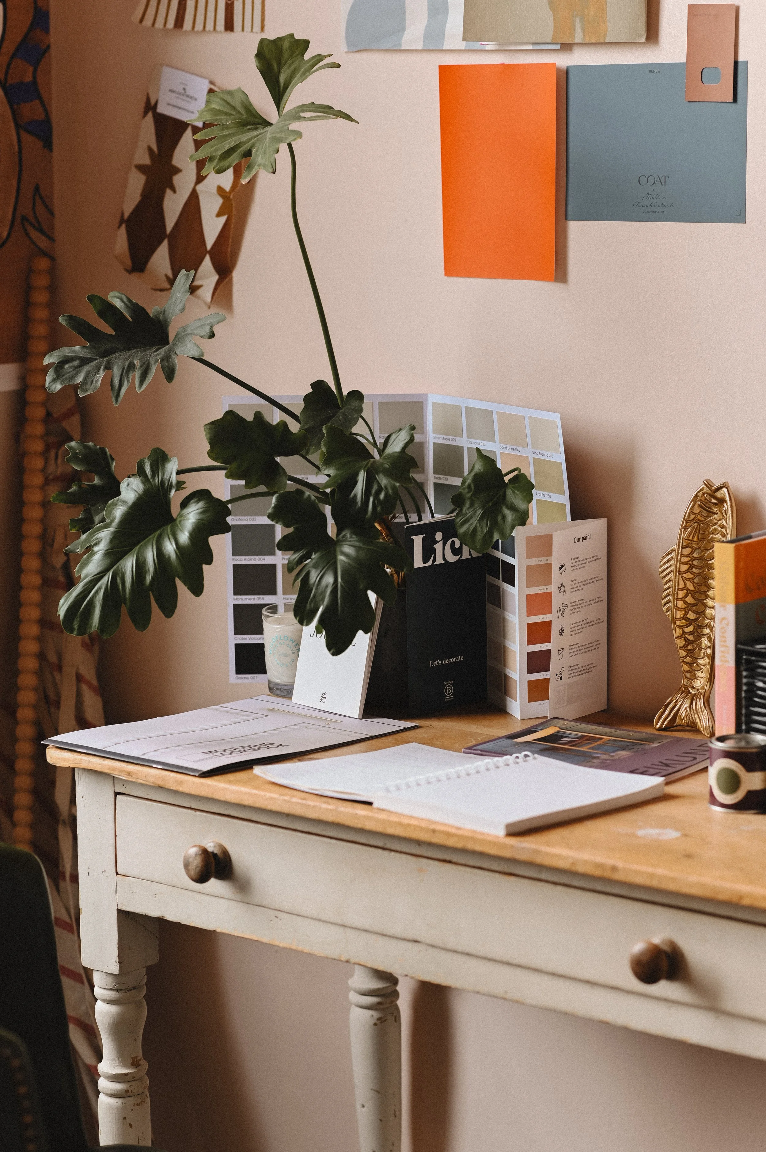 A cluttered workspace with a small, white distressed table holding open notebooks, a poster with color swatches, and a potted plant. The background features a wall with hanging paper samples or notes and a decorative gold-colored figurine of a fish.