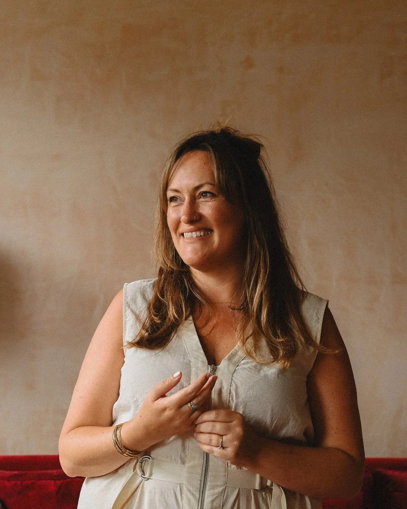 An interior designer with long wavy hair wearing a sleeveless, light-colored dress, standing against a plain, textured beige wall.