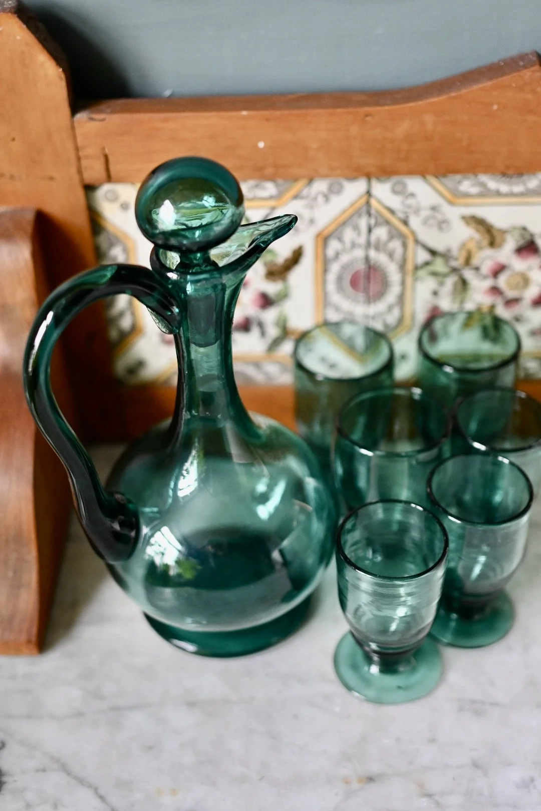 Green glass decanter with a round stopper and six matching glasses on a marble surface, with a wooden chair and patterned cushion in the background.