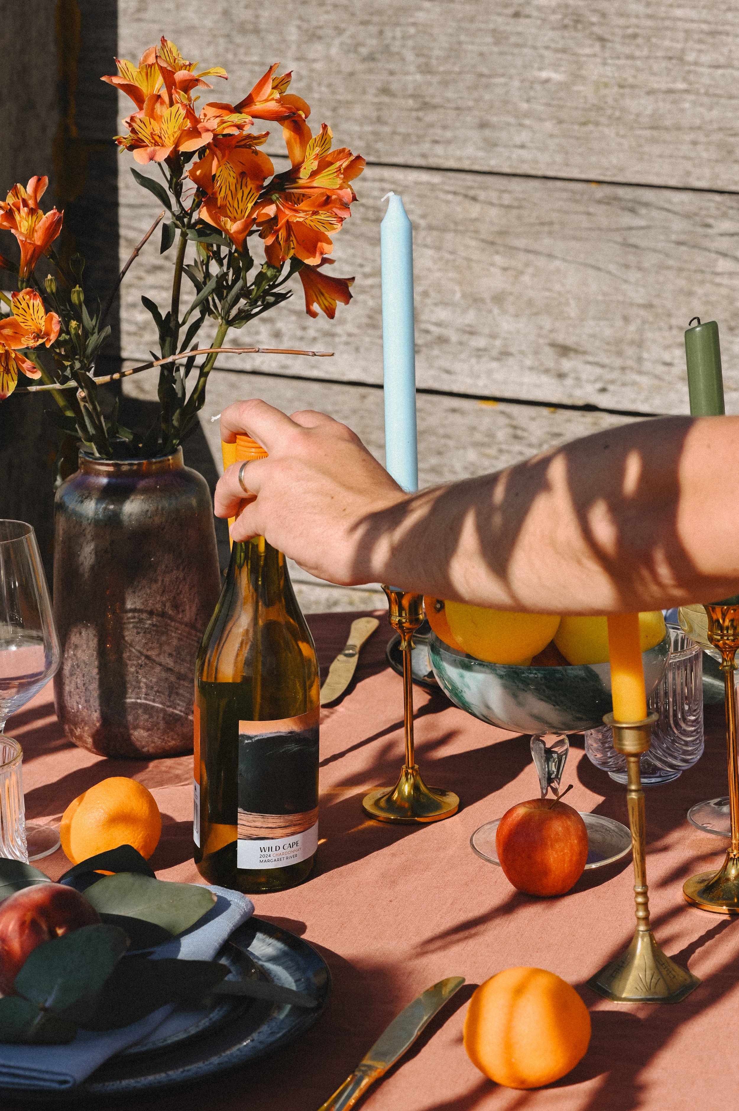 A table set with flowers, candles, fruits, and wine bottles. The flowers are in a brown vase, and there are multiple candles in gold holders. The tablecloth is pink, and the background is a wooden surface. There are oranges, peaches, and a bowl of lemons on the table, along with some cutlery and plates.