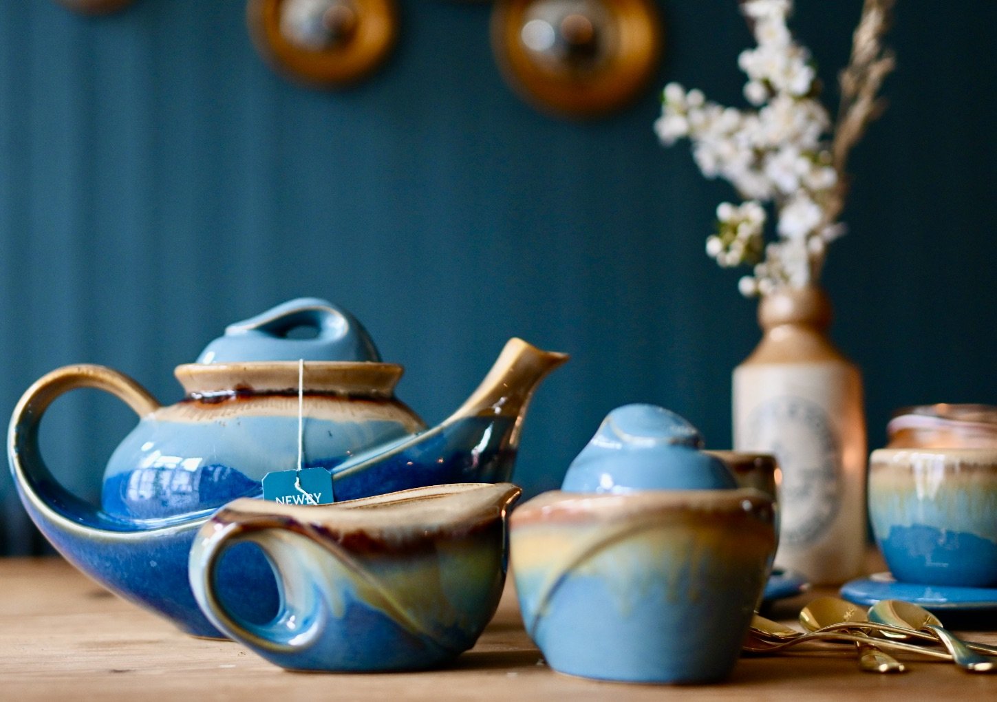 Set of blue and brown ceramic teapots and cups on a wooden table, with a vase of white flowers in the background.
