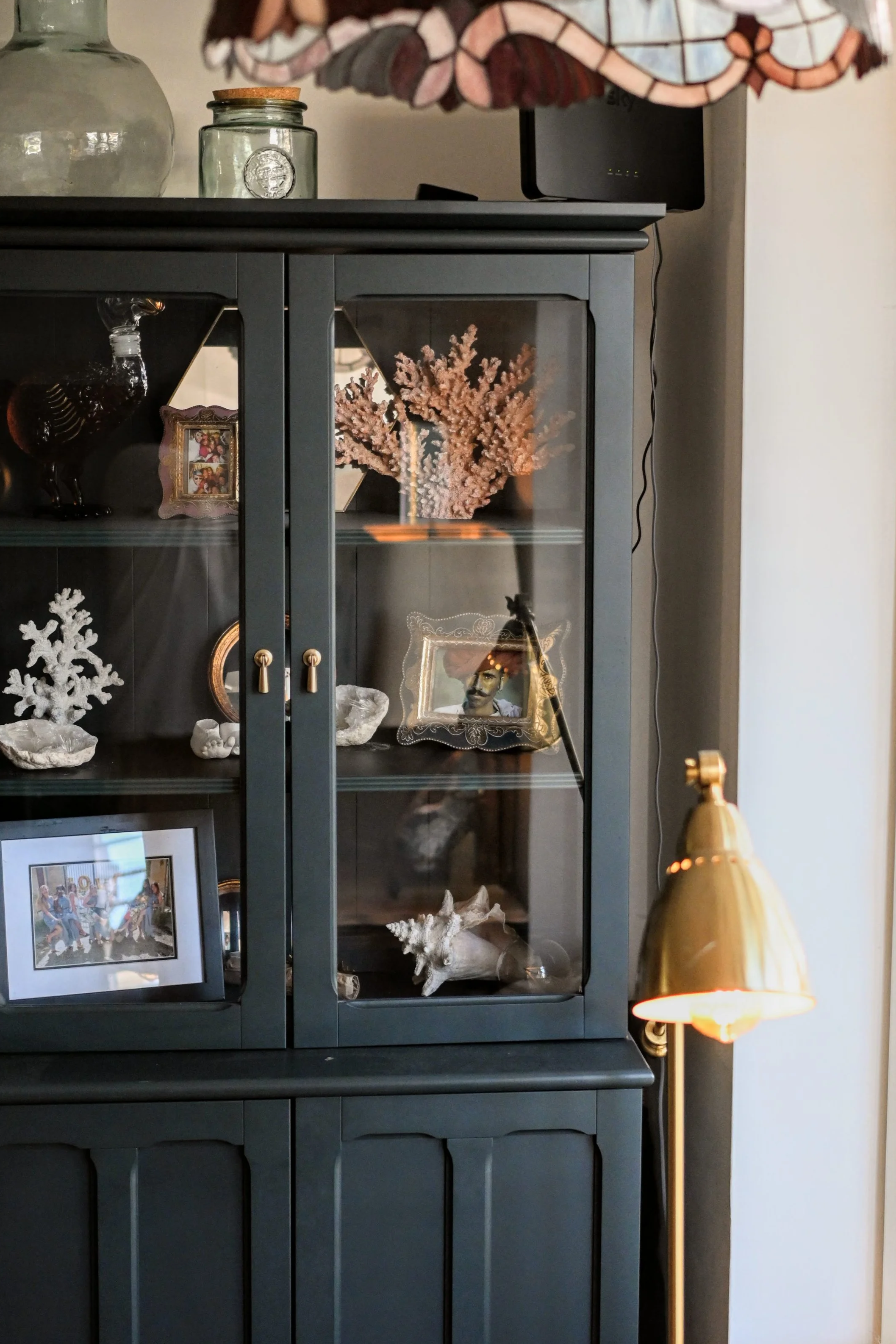 A dark blue display cabinet filled with decorative items such as framed photos, coral arrangements, white coral sculptures, and glass figurines. On top of the cabinet, there are jars and a stained glass lampshade. To the right of the cabinet, a gold-colored floor lamp with a pointed shade is visible.
