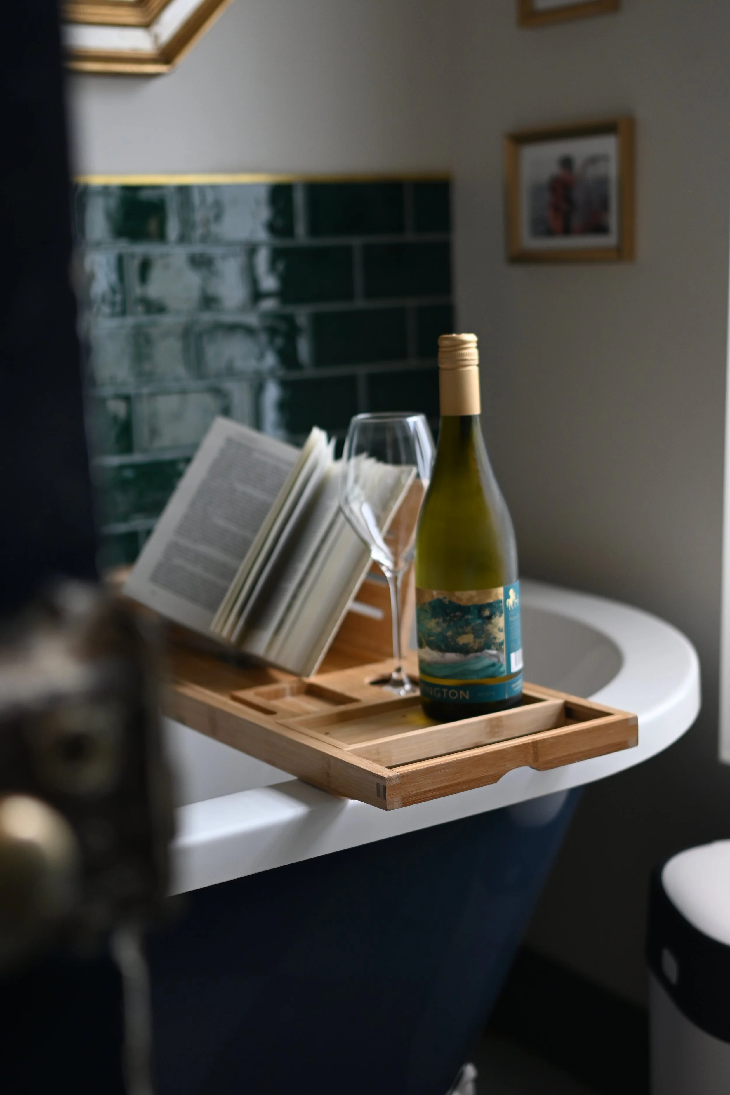 A wooden tray on a white table holds a bottle of white wine, a wine glass, and an open book. In the background, there is a picture frame on the wall and part of a brick wall. The scene appears to be a cozy indoor setting.