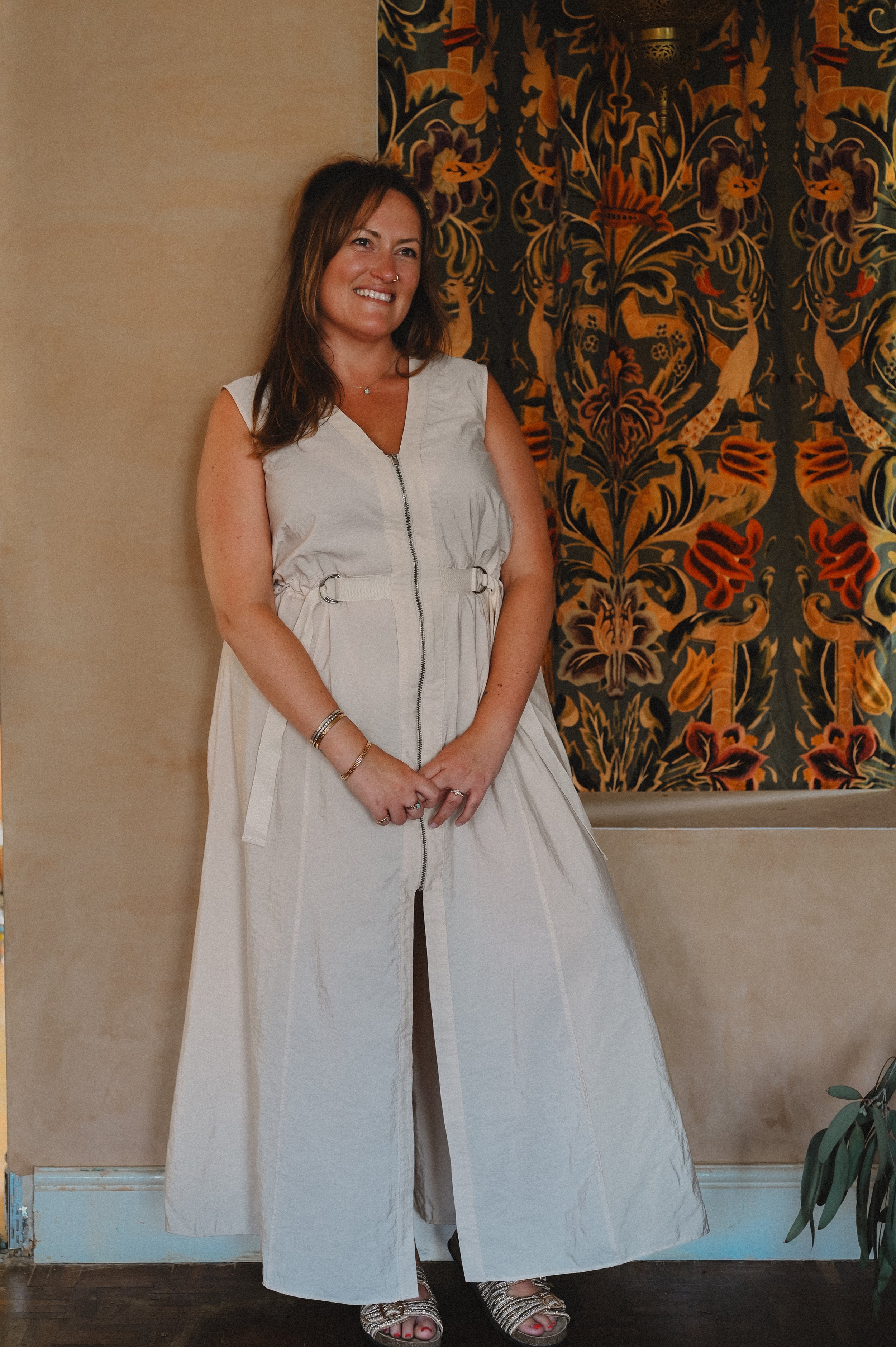 A woman with shoulder-length brown hair, wearing a sleeveless white zip-up dress and silver sandals, standing in front of a decorative tapestry with floral and peacock designs, smiling at the camera.