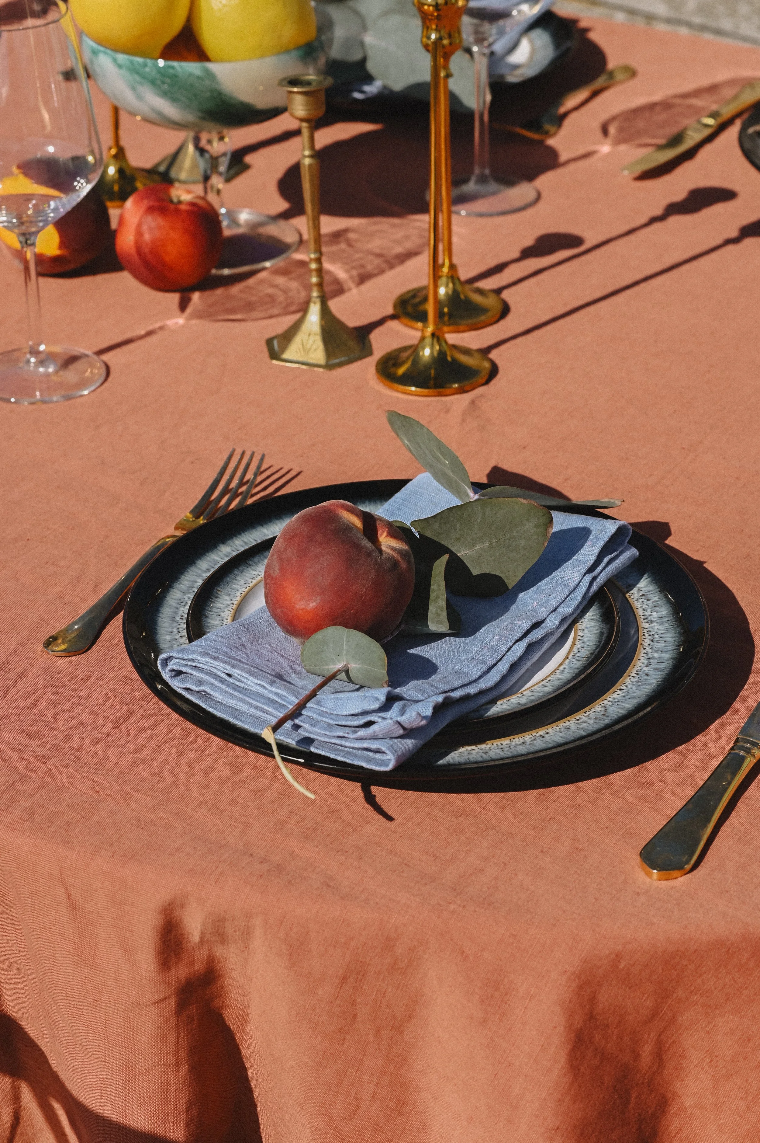 Outdoor table setting with a black plate, silver fork, and blue cloth napkin. Two peaches with green leaves on the napkin. In the background, a pink tablecloth with gold candlesticks, a bowl of fruit, and a glass of water.