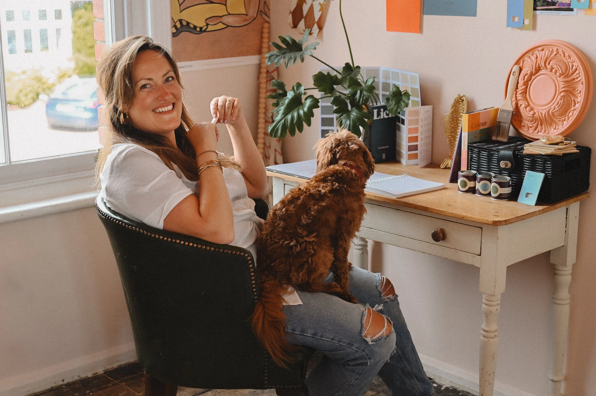 A woman sitting in a black chair at a desk with a small dog on her lap, smiling at the camera inside a room decorated with colorful artwork, plants, and various items on the desk.