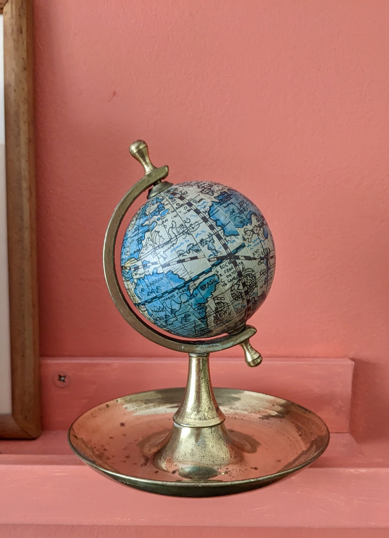 A globe with a vintage brass stand on a wooden surface against a pink wall background.