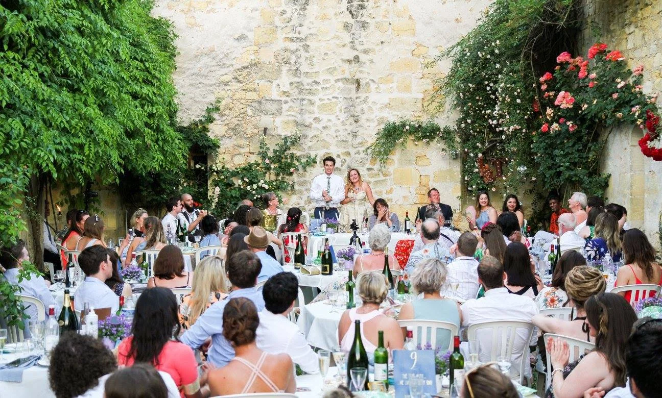 A wedding reception outdoors with guests seated at round tables, a bride and groom standing at a head table, surrounded by greenery and colorful flowers.