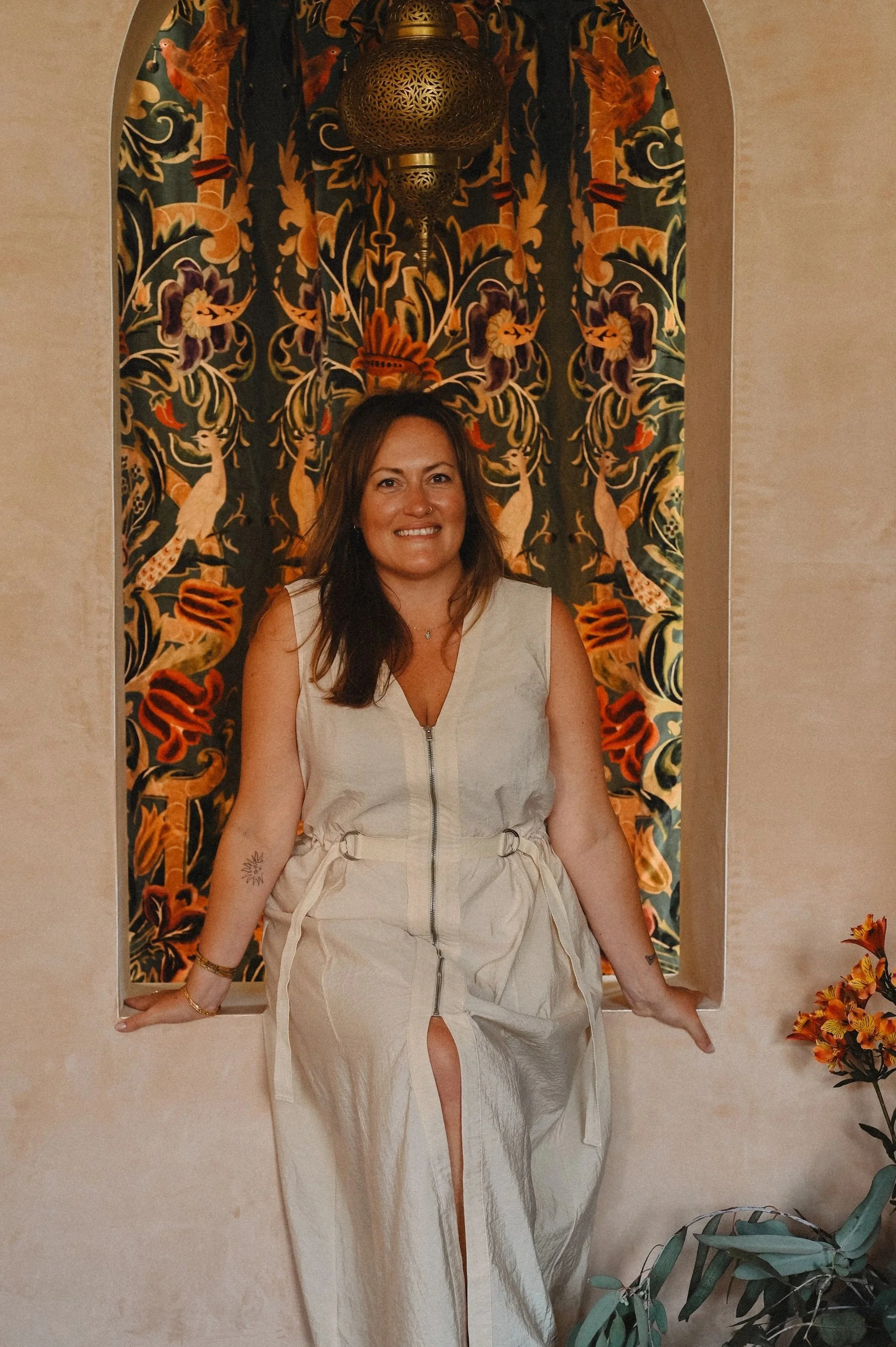 A woman with long brown hair and a tan complexion smiling, standing in front of a colorful tapestry with bird and floral patterns. She is wearing a sleeveless beige dress with a zipper and drawstring waist, and a wrist bracelet. There are orange flowers on the right side and green foliage on the bottom right in the foreground.