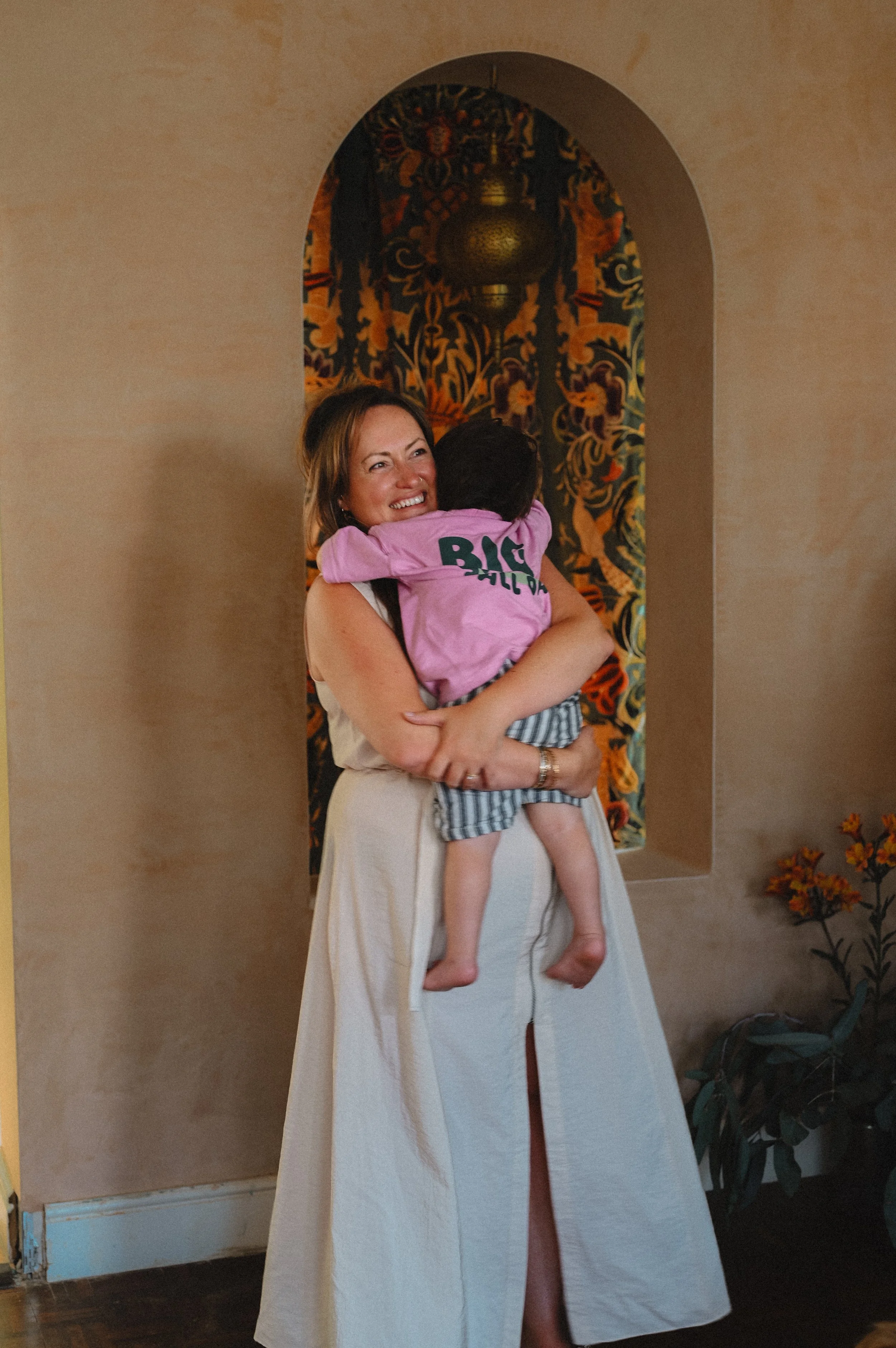 A woman with shoulder-length hair is smiling and holding a young child who is wearing a pink shirt and striped shorts. The woman is wearing a light-colored skirt with a slit. They are standing in a room with a decorative painted niche containing a hanging ornament, against a beige wall with some plants visible on the side.