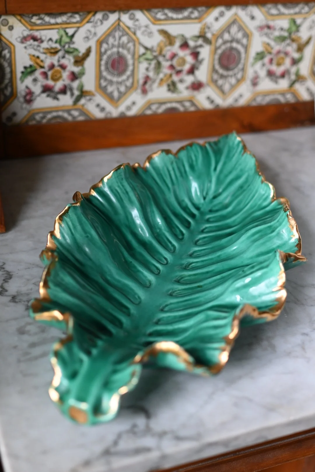 A decorative green ceramic dish shaped like a leaf with gold trim, placed on a marble surface in front of a tiled wall with floral and geometric patterns.