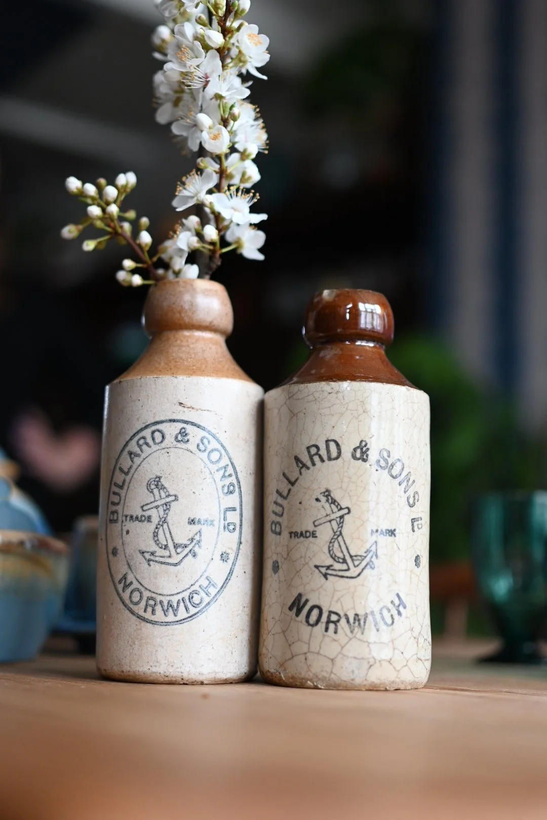Two vintage ceramic bottles with 'BULLARD & SONS, NORWICH' and an anchor logo, one holding a sprig of white flowers, on a wooden surface.