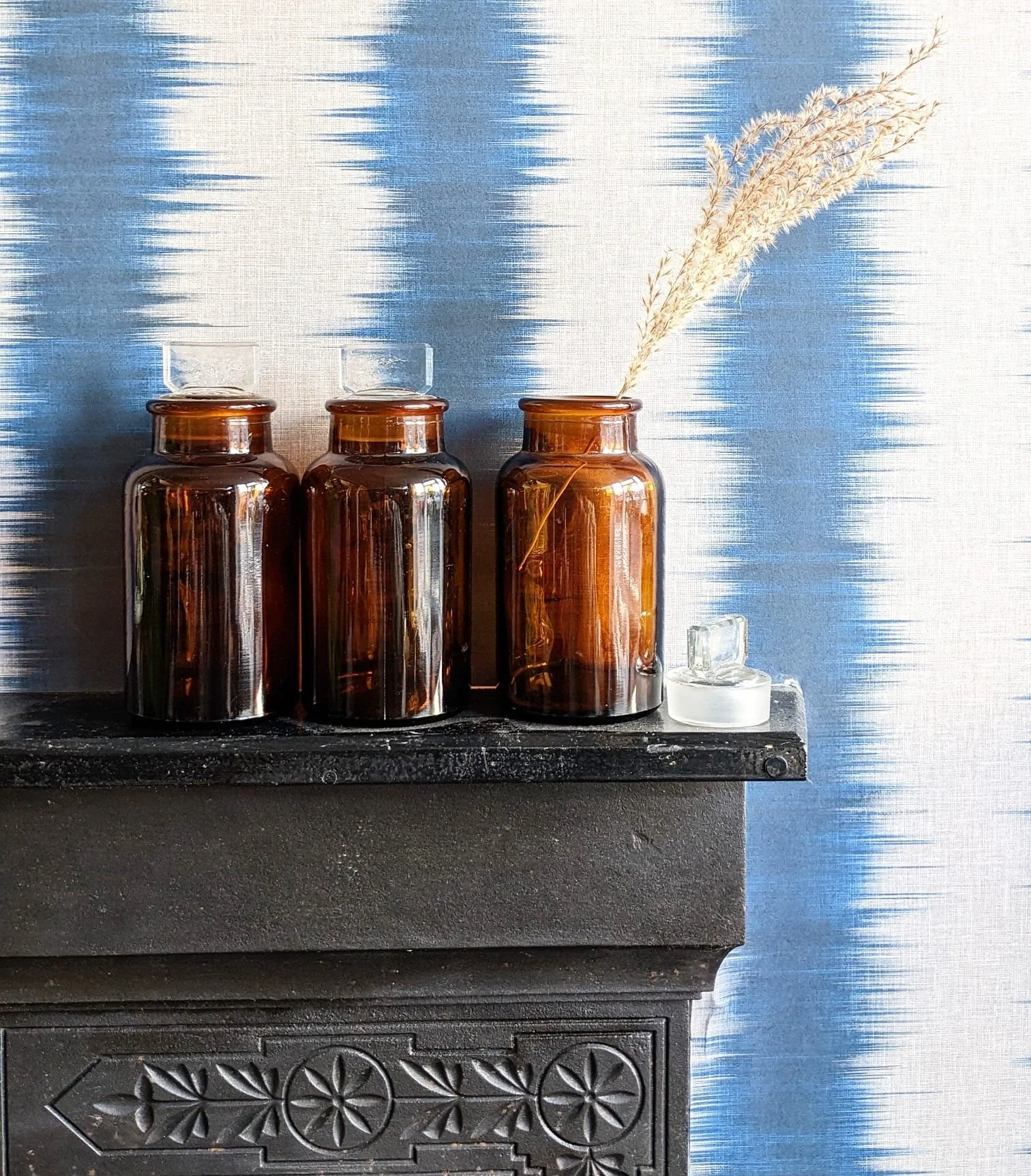 Three brown glass bottles with glass stoppers on a black textured surface, next to a small glass container with a white lid. One bottle has dried wheat stalks inside. The background features a blue and white textured wall.