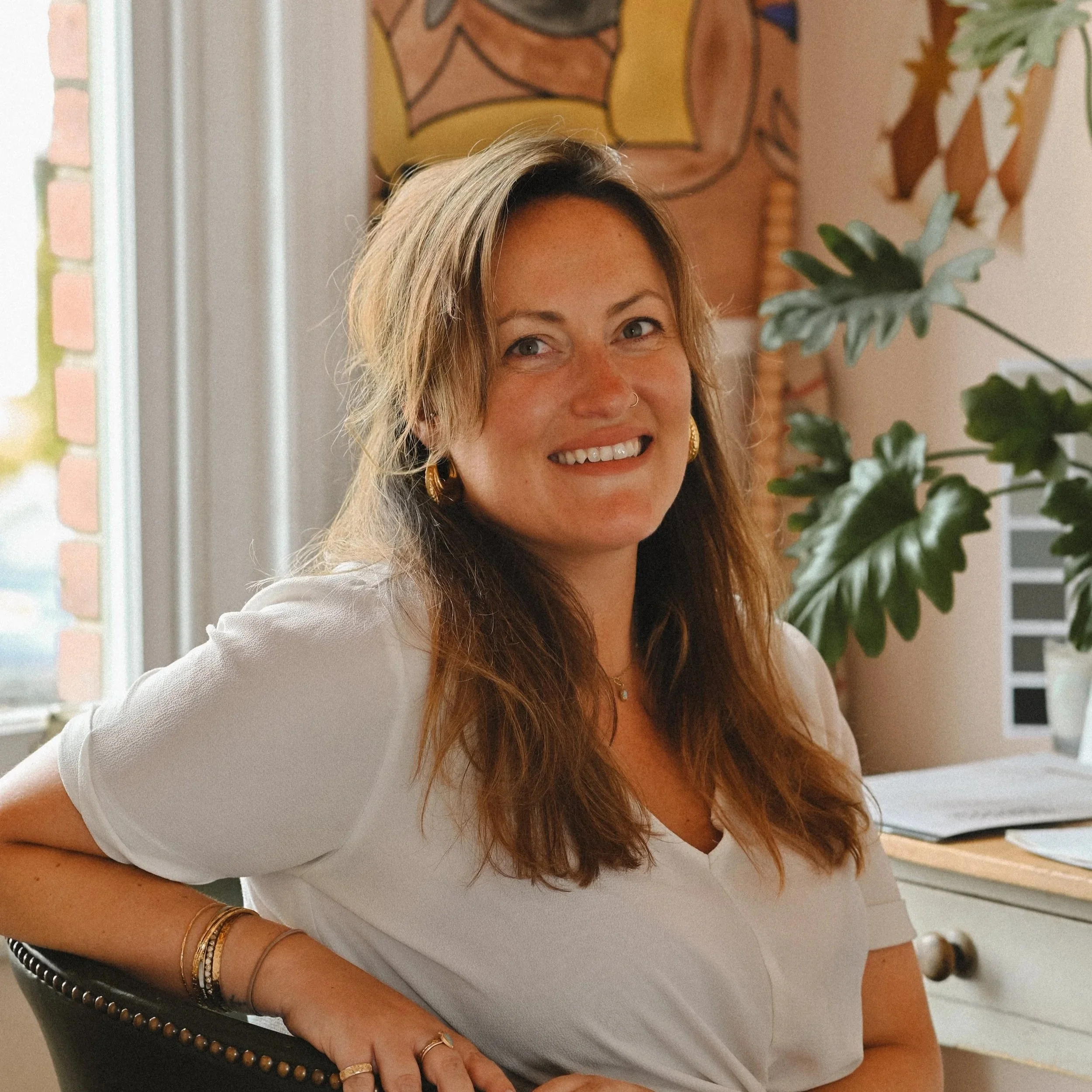 Close-up of a smiling woman with long hair, wearing a white top, gold earrings, and bracelets, sitting in a room decorated with art and plants.