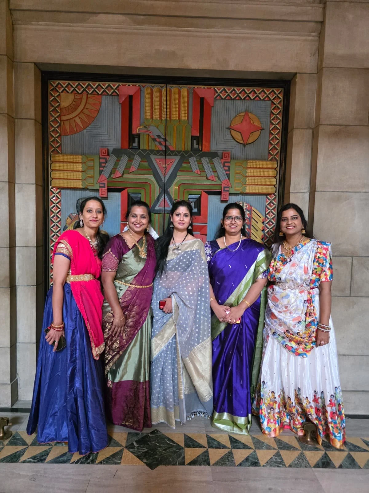 Five women in colorful traditional Indian sarees and dresses standing together in front of an ornate, geometric wall art.
