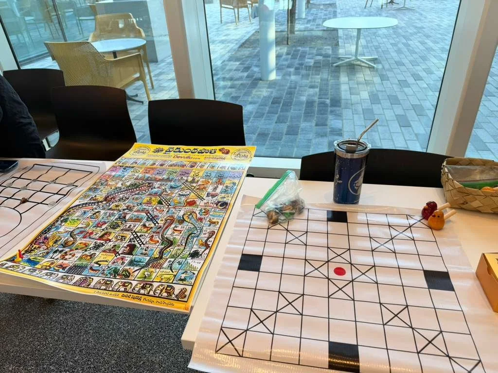 Table with a colorful board game, a tic-tac-toe grid, a soda with a straw, and some small toys, set in front of large windows showing outdoor patio furniture.