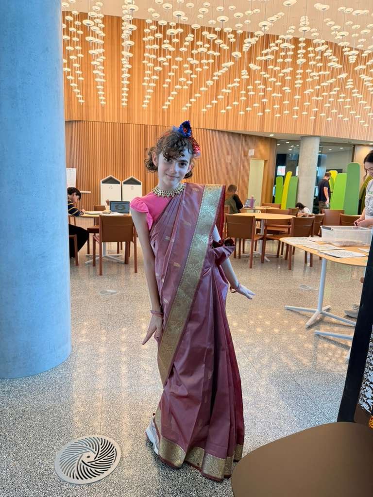 A young girl dressed in a pink and gold traditional Indian sari, accessorized with a necklace and a blue hair ornament, standing in a modern indoor space with wooden walls, ceiling light fixtures, and people seated at tables.