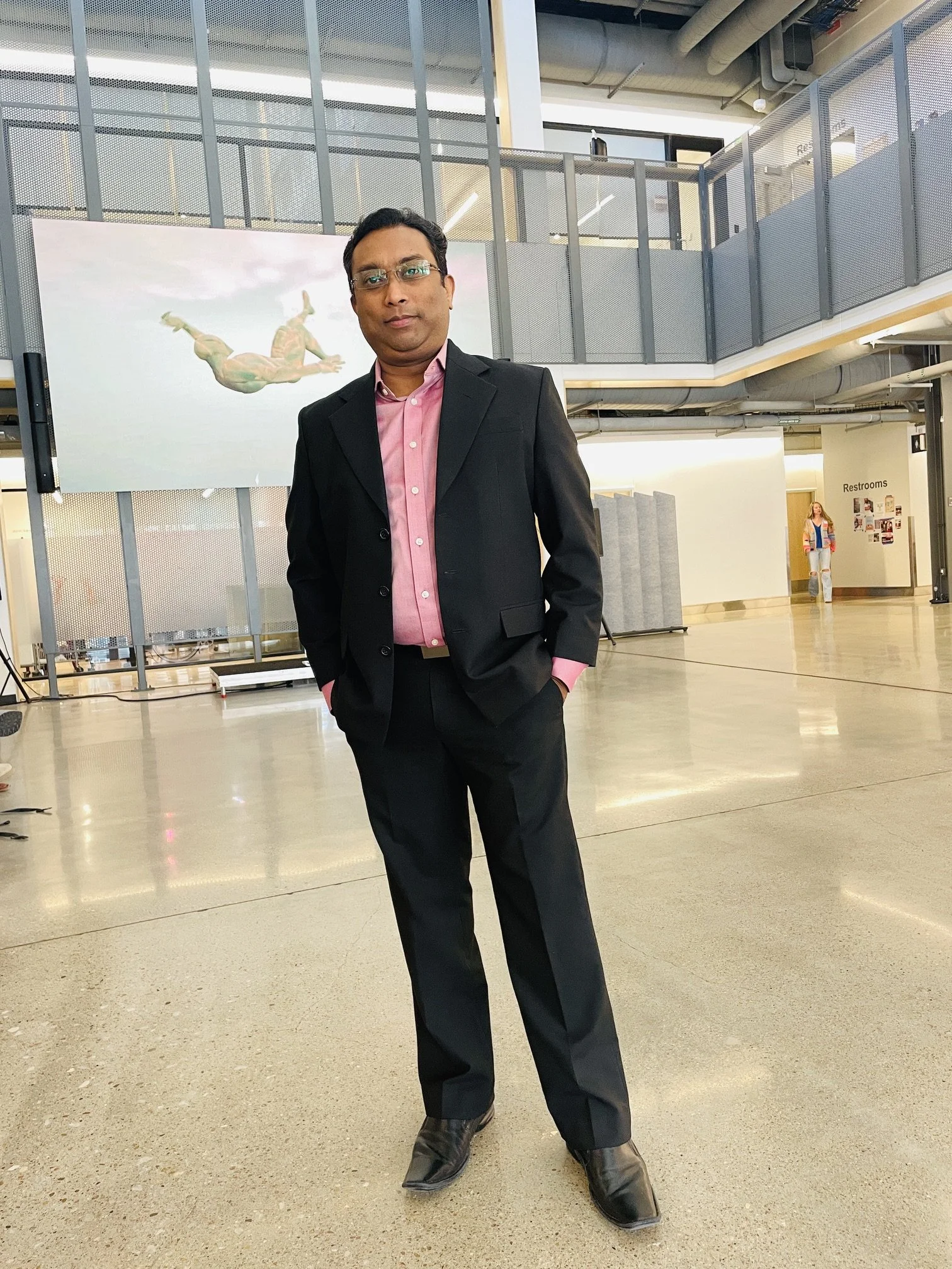 A man wearing a black suit and pink shirt standing in an indoor space with a person walking in the background near a restroom sign, and an animated diver sticker on the wall.