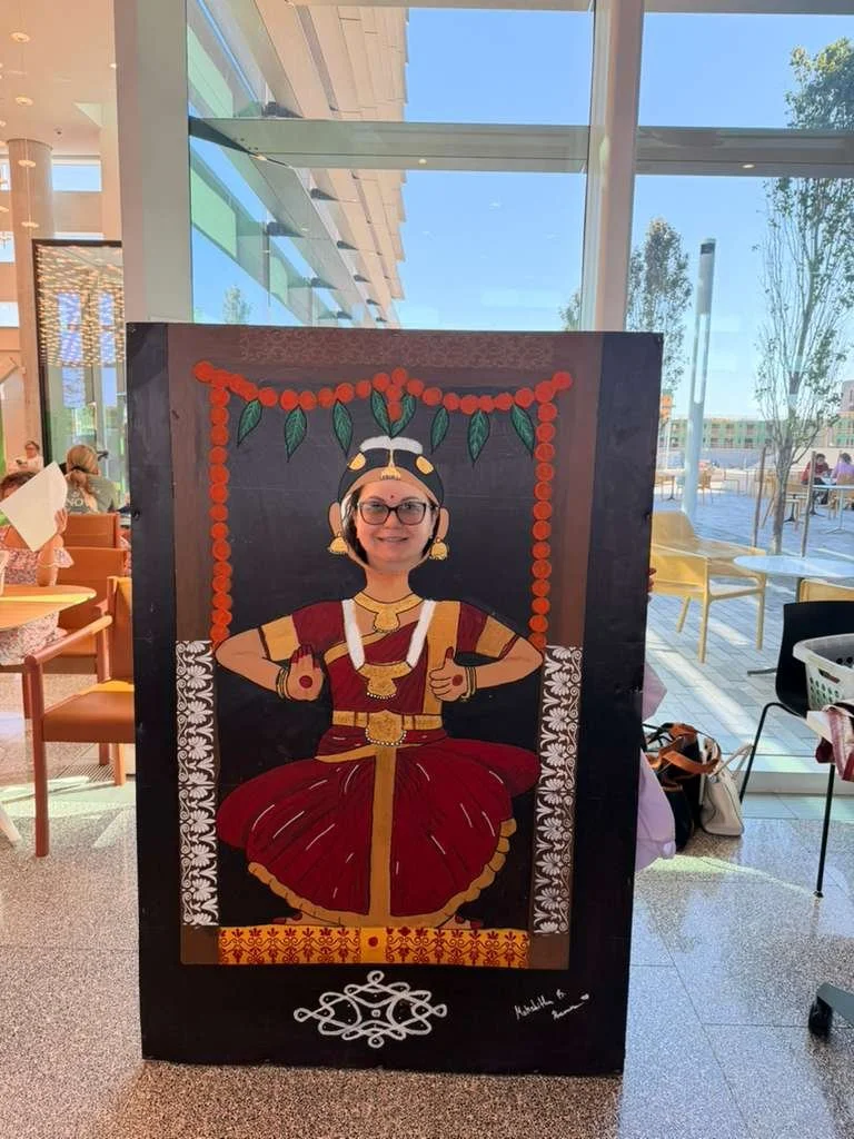 Person posing behind a painted cutout board with a traditional Indian dancer illustration, only their face is visible, wearing glasses and a headband.