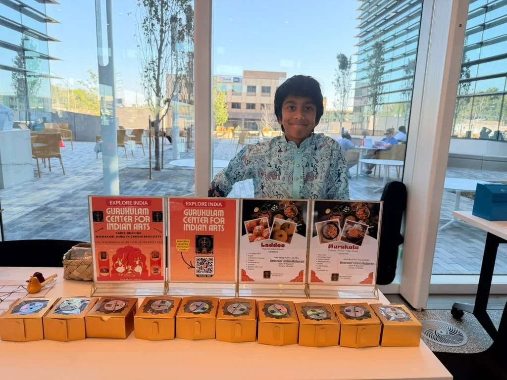 A young boy standing behind a table with Indian arts and food promotion materials, in a bright indoor space with large glass windows showing an outdoor patio and modern buildings.