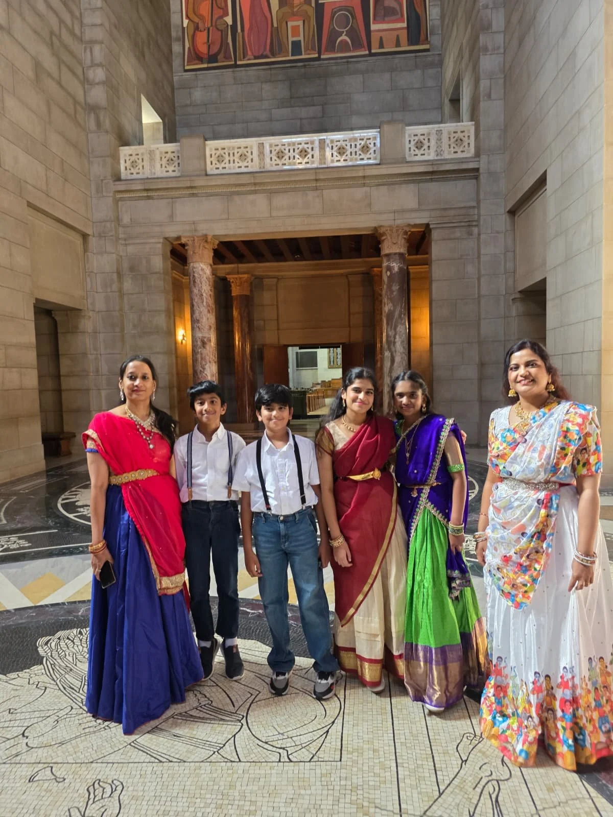 A group of six people, including four women and two young boys, standing inside a large, elegant building with stone walls and decorative columns. The women are dressed in colorful traditional Indian sarees, and the boys are wearing white shirts with