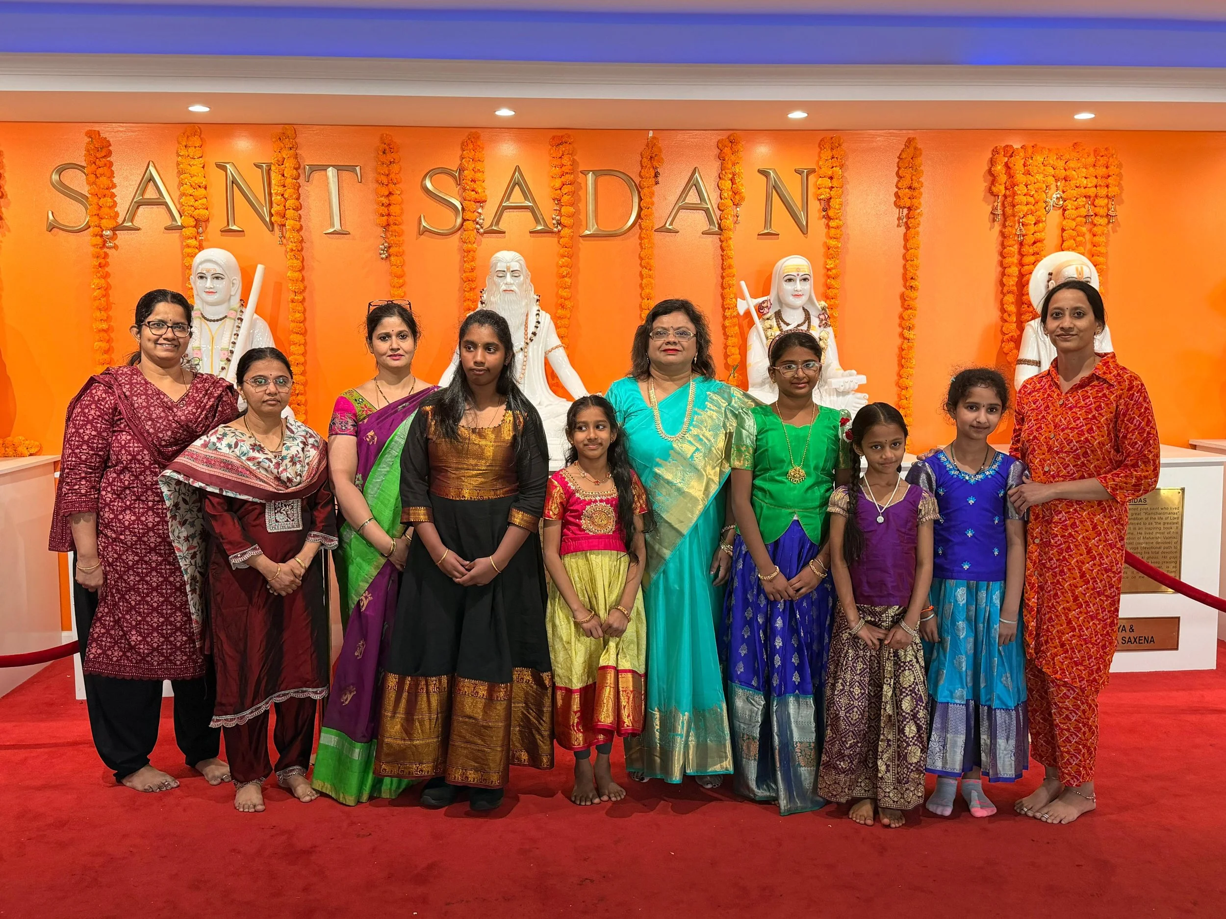 Group of women and children dressed in traditional Indian sarees, standing in front of orange backdrop decorated with marigold flower garlands and Hindu deity sculptures, with the partial text 'SANT SADAN' visible in the background.