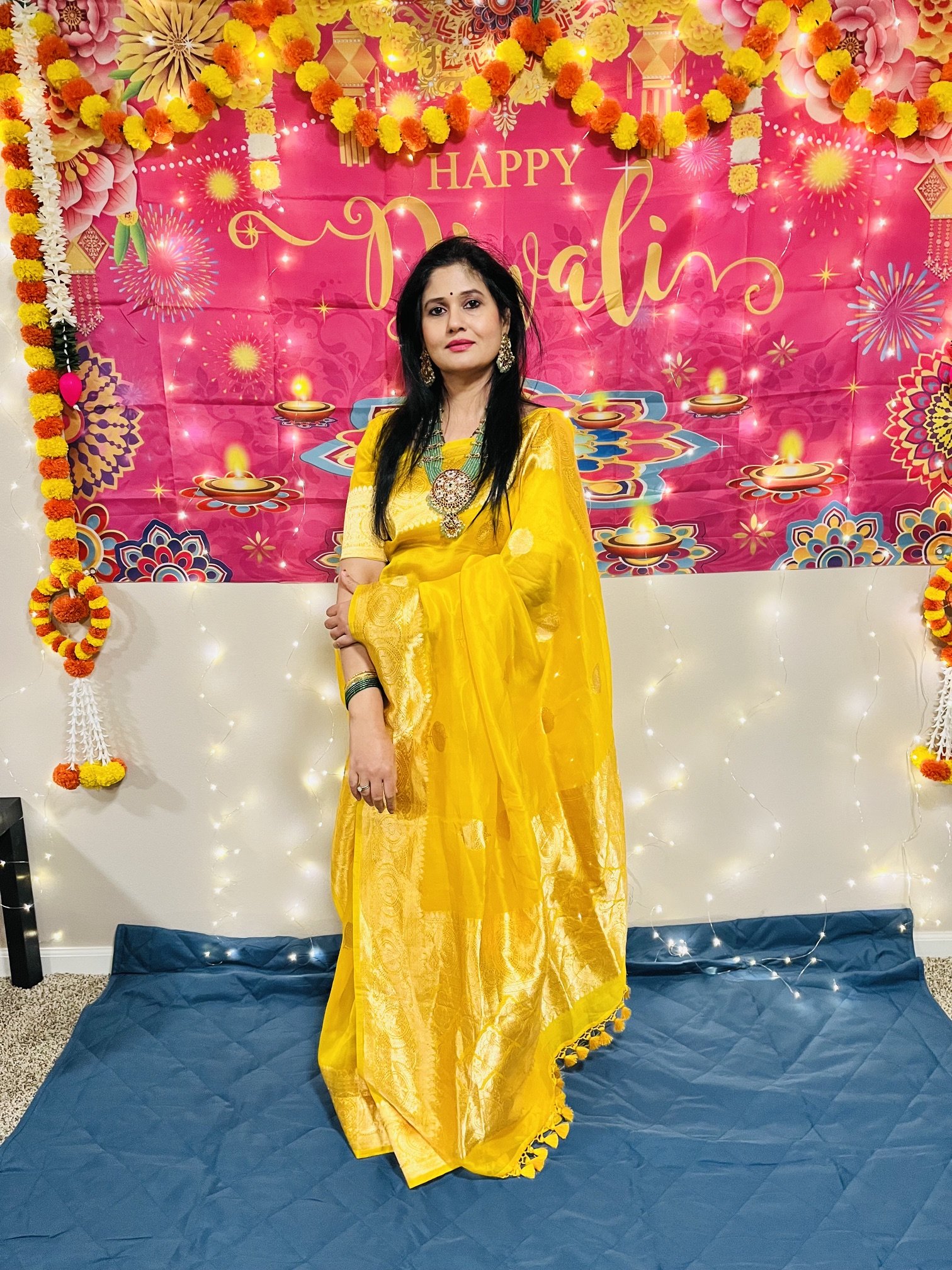 A woman dressed in a yellow saree standing in front of a pink decorated backdrop with the words "Happy Diwali" and lit oil lamps. The backdrop features flowers, fireworks, and fairy lights, with marigold flower garlands on the sides.