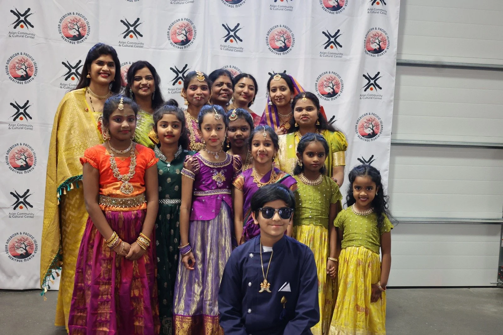 Group of women and children dressed in colorful traditional Indian attire standing in front of a banner with the Asian American & Pacific Islander Heritage Month logo.