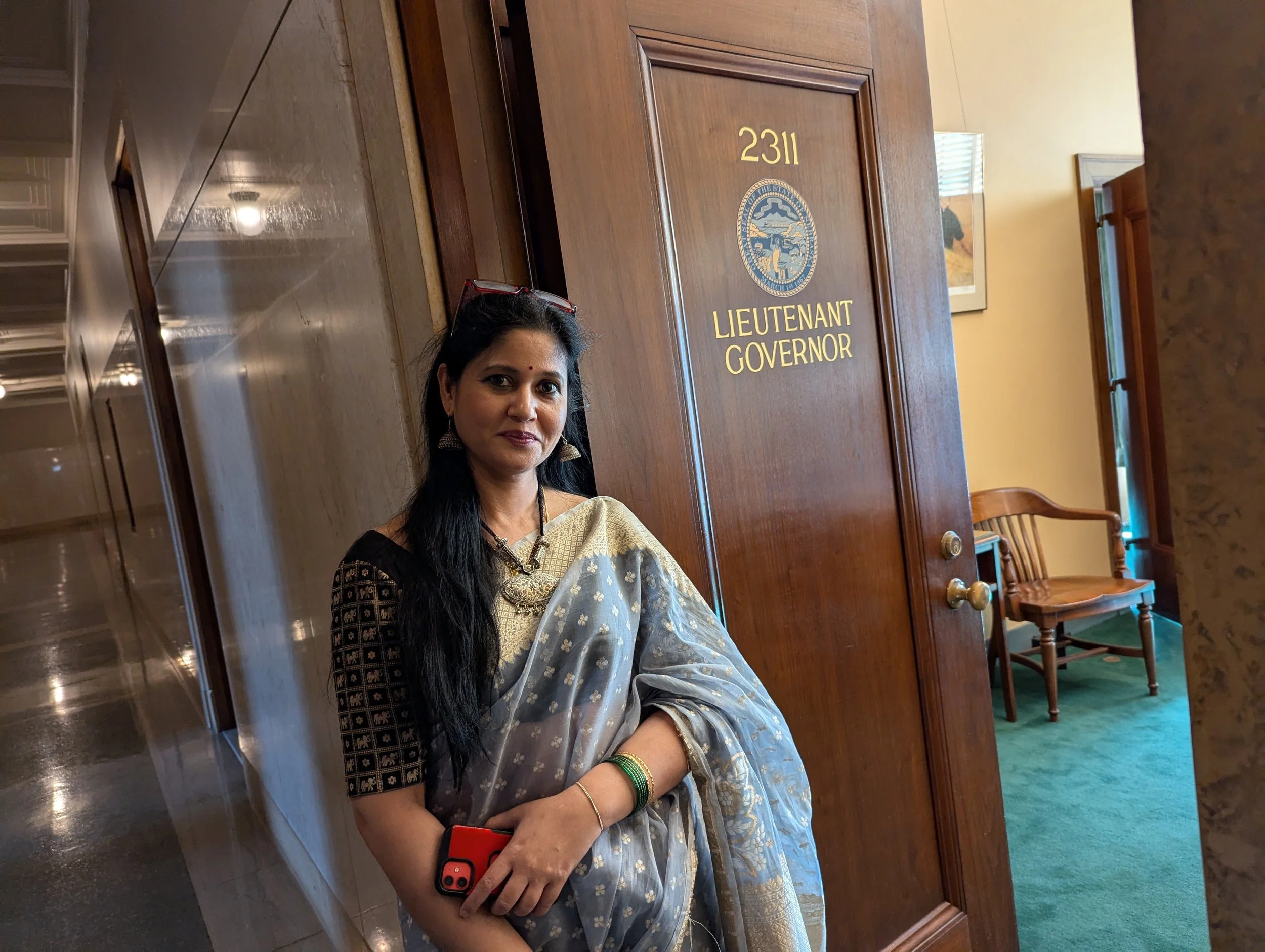 A woman with long black hair, wearing traditional Indian attire with a cream-colored sari and gold jewelry, is standing in front of a wooden door labeled '2311 Lieutenant Governor' with an official seal. She is holding a phone in her left hand and ha