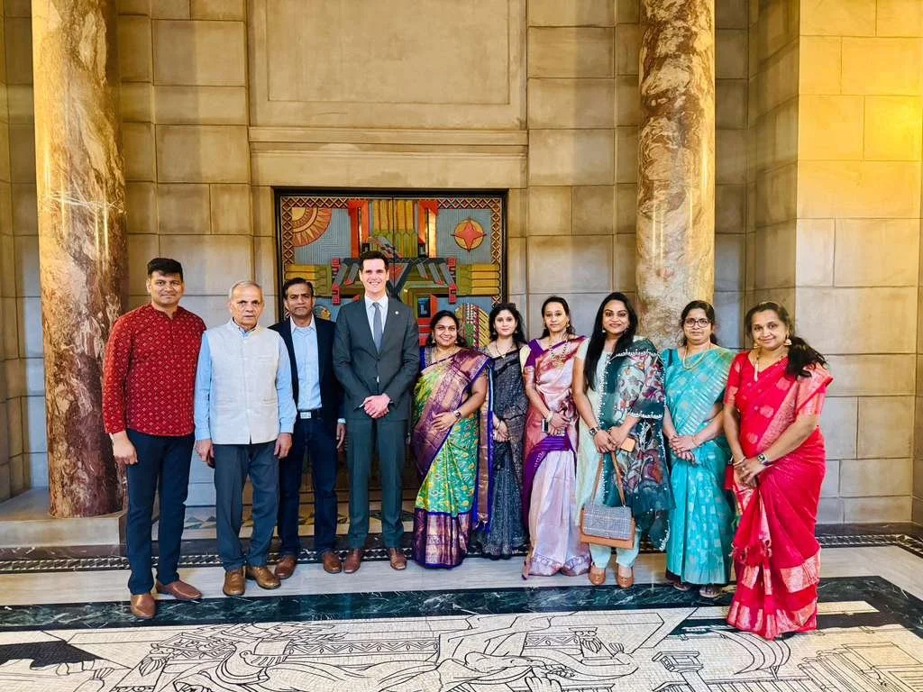 Group of ten people standing in front of a decorative wall with artwork, dressed in a mix of traditional Indian and Western clothing.