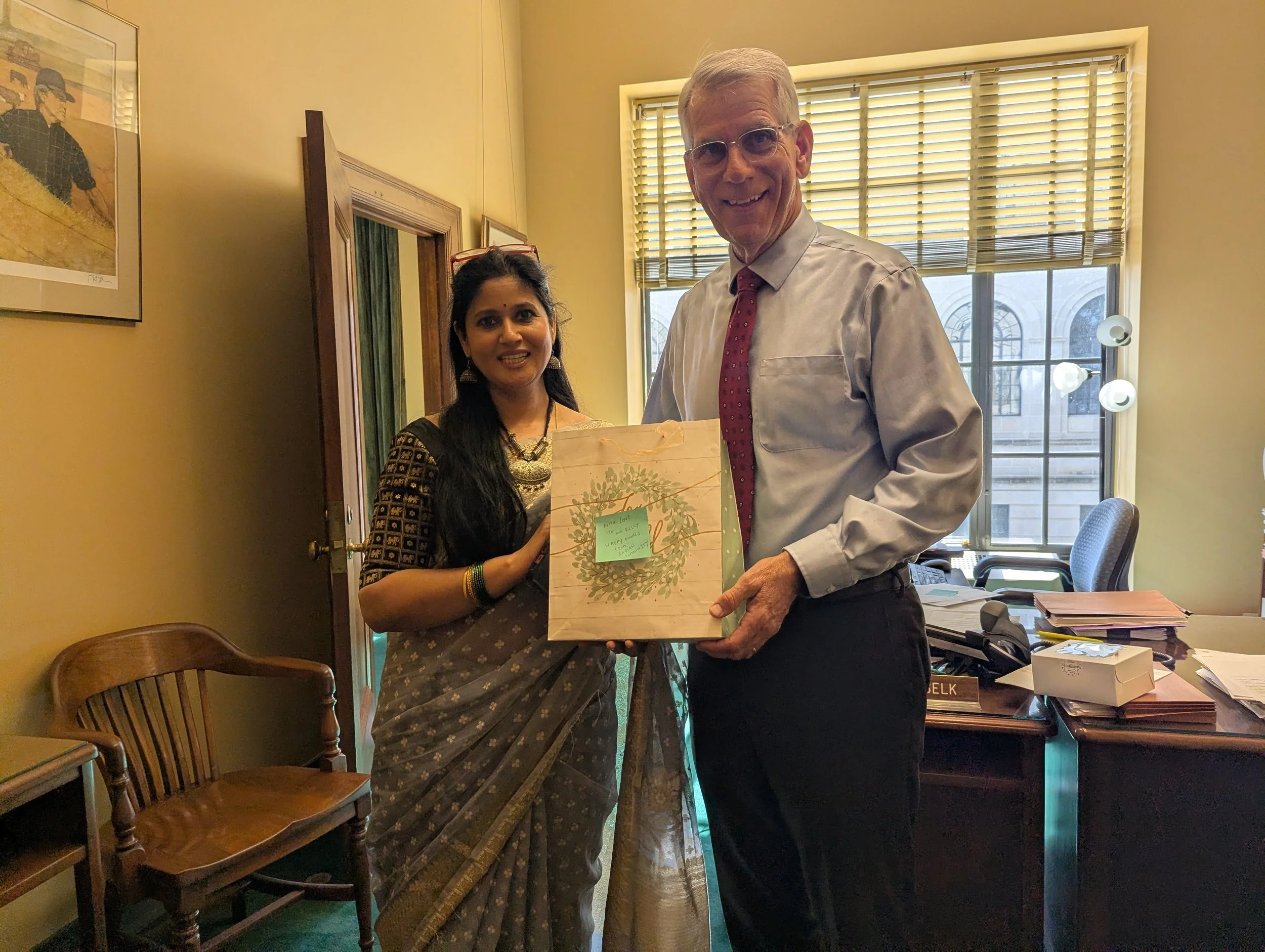Two people standing in a room. The woman is wearing traditional Indian attire and glasses, holding a gift bag with botanical design. The man is dressed in a shirt and tie, smiling. There are chairs, a desk with papers, and large windows with horizont
