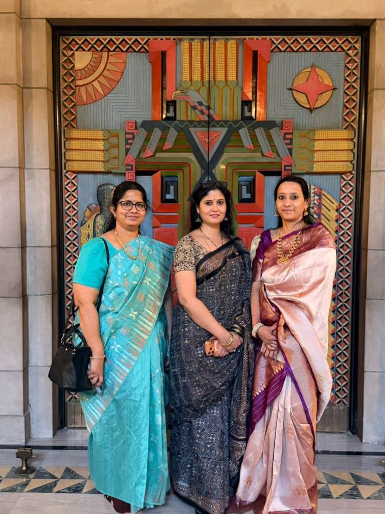 Three women dressed in traditional Indian sarees standing together in front of colorful geometric artwork.
