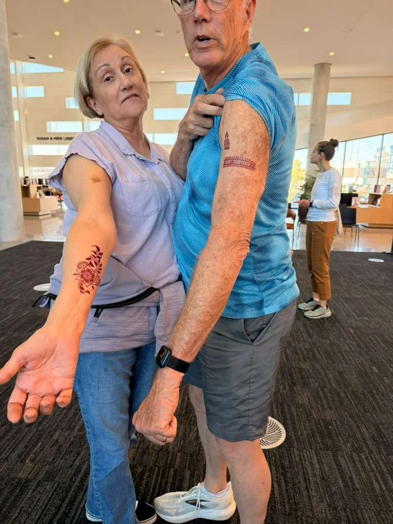 An elderly couple displaying tattoos on their upper arms inside a modern, well-lit building, possibly an airport or lobby, with a woman in the background looking away.