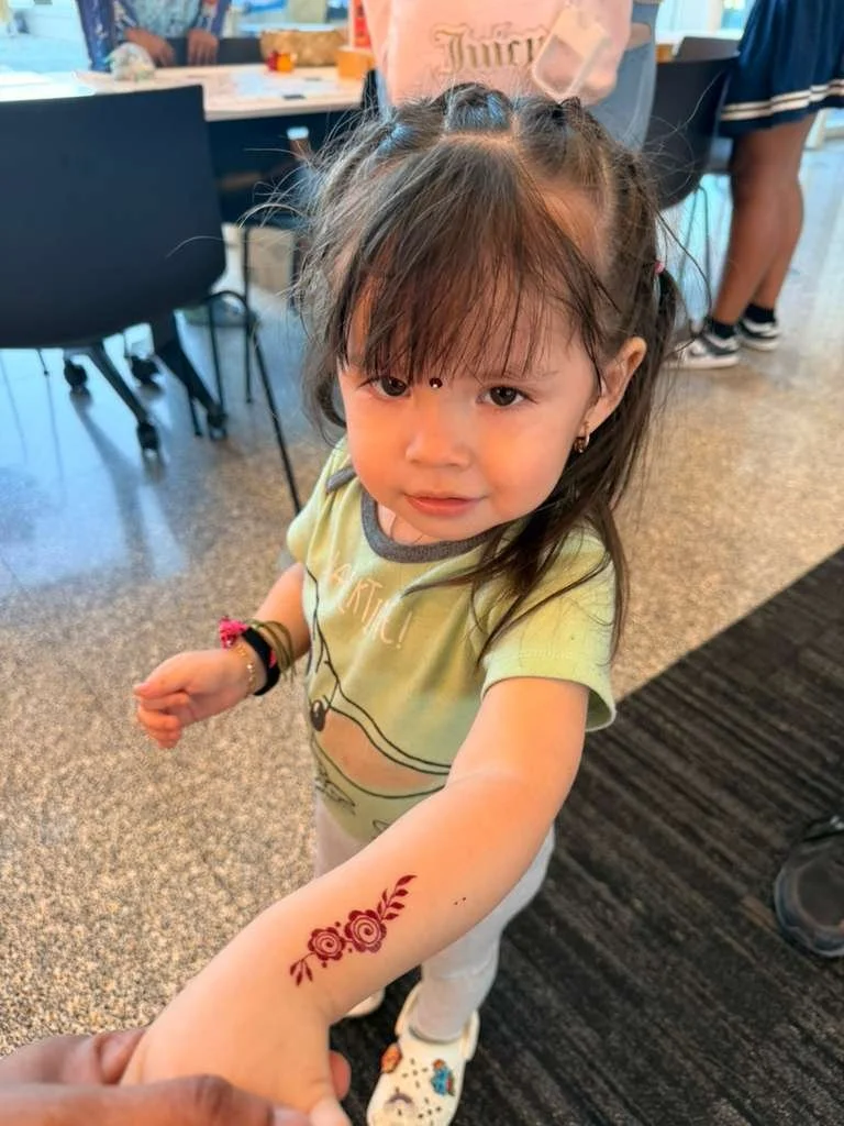 A young girl showing her arm with a henna tattoo of red roses and leaves, inside a busy indoor space with chairs and people in the background.