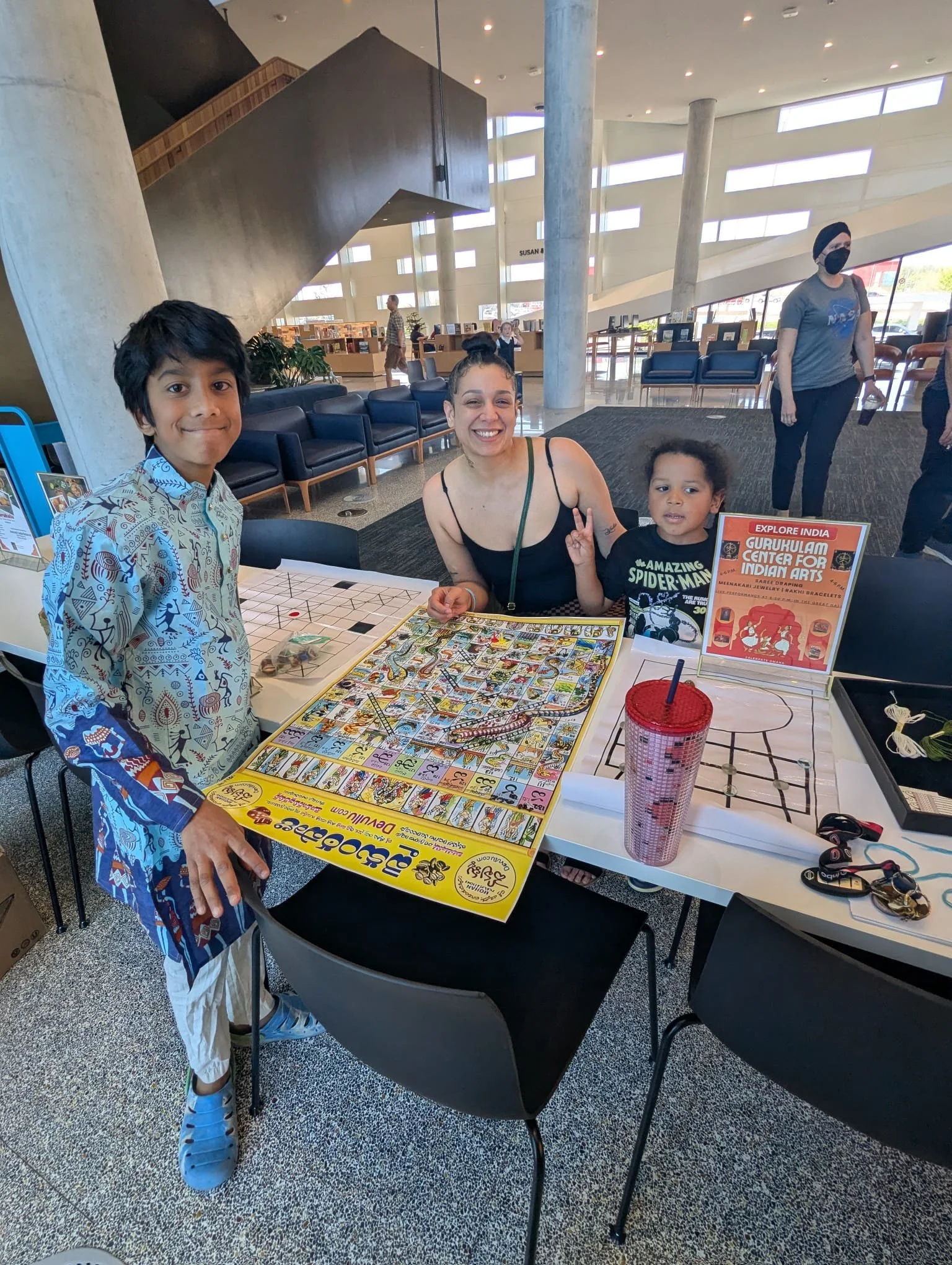Group of four people, including two children and two adults, sitting and standing around a table with a colorful board game at a public space with modern architecture and bookshelves in the background.