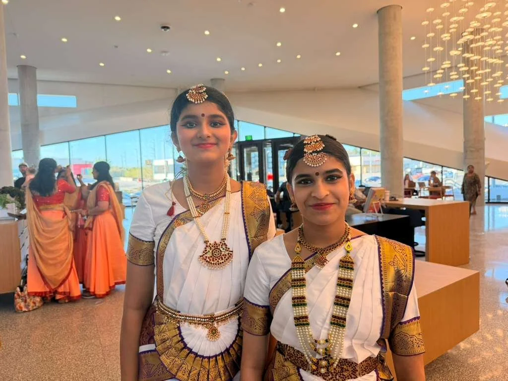 Two young girls wearing traditional Indian sarees and jewelry stand inside a modern, well-lit building, with other women in sarees in the background.